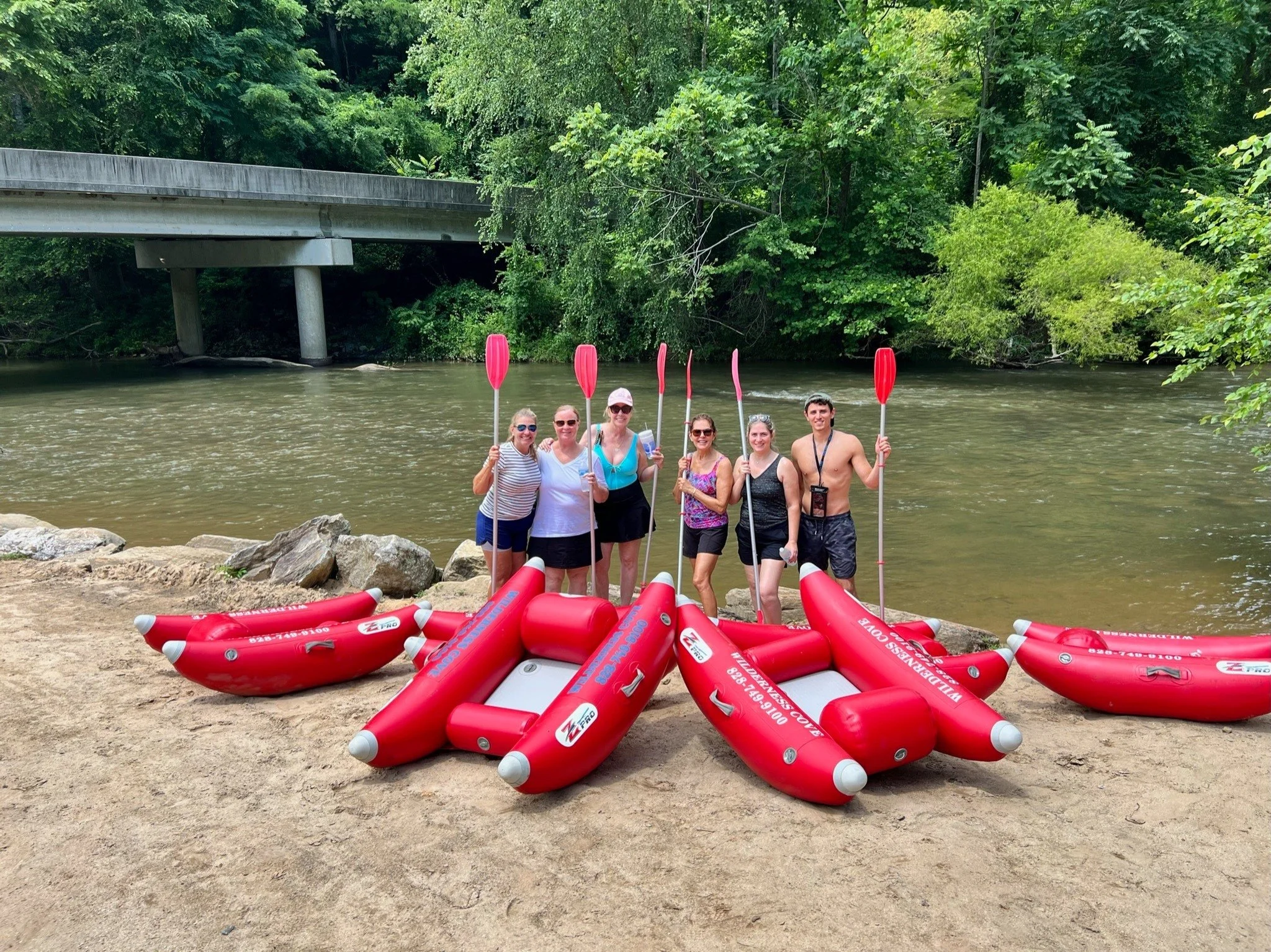 A group of six people standing by a river holding paddles, with a pile of red inflatable boats on the sandy riverbank in front of them, a bridge crossing over the river, and lush green trees in the background.