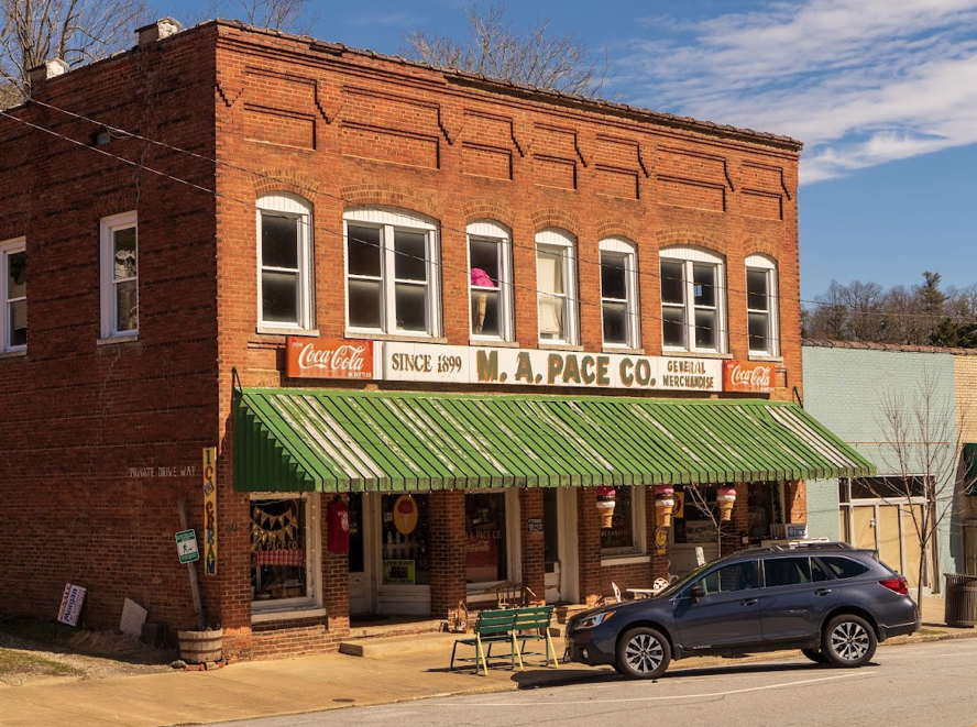 A two-story red brick building with a green striped awning, featuring a sign for M. A. Pace Co., established 1899, with Coca-Cola signage. There is a black SUV parked in front and a sidewalk with a bench and some signs, under a partly cloudy sky.