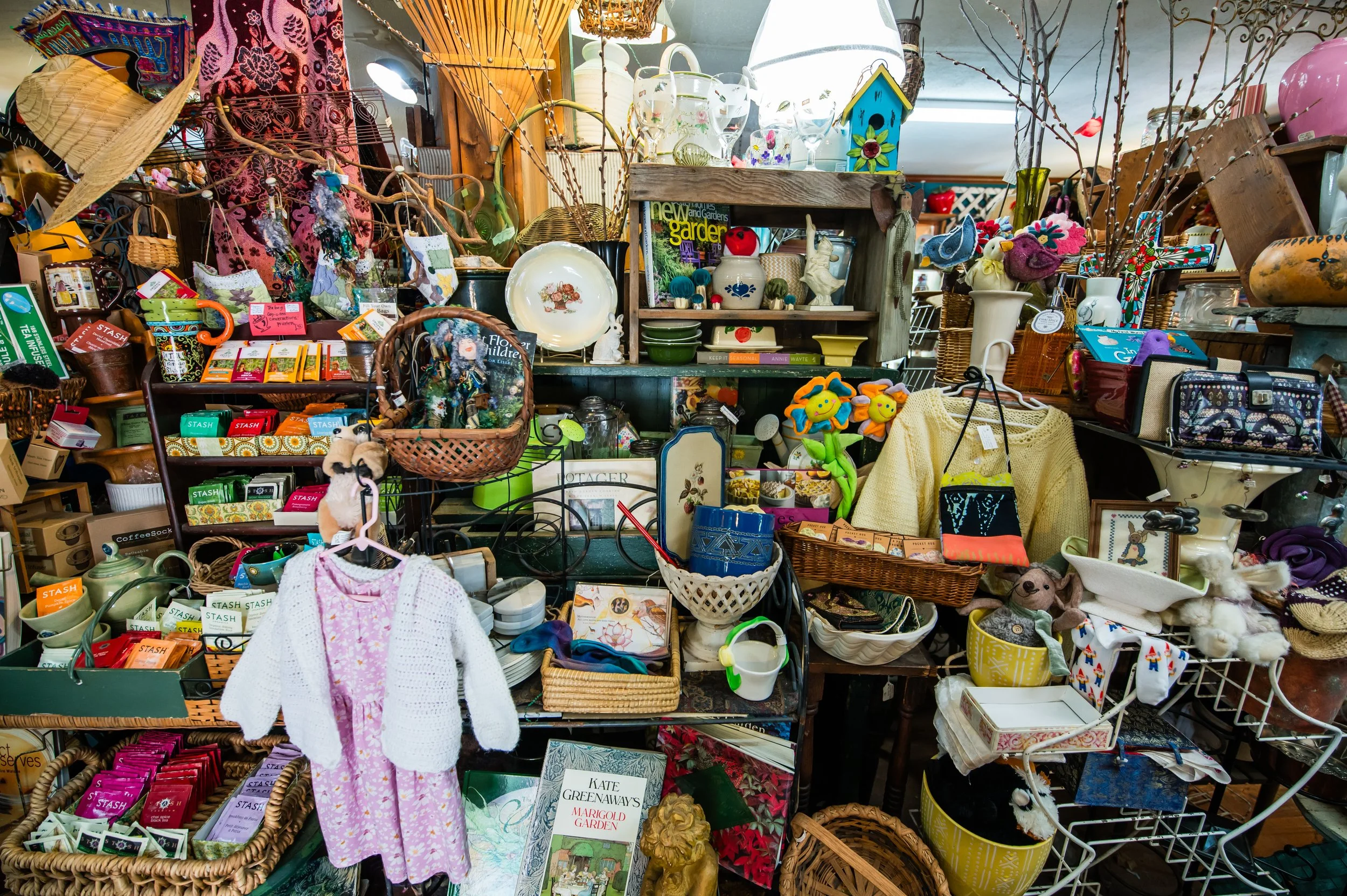 A cluttered display of various household items including kitchenware, decorative objects, plants, toys, and clothing all arranged on shelves and tables at a thrift store or flea market.