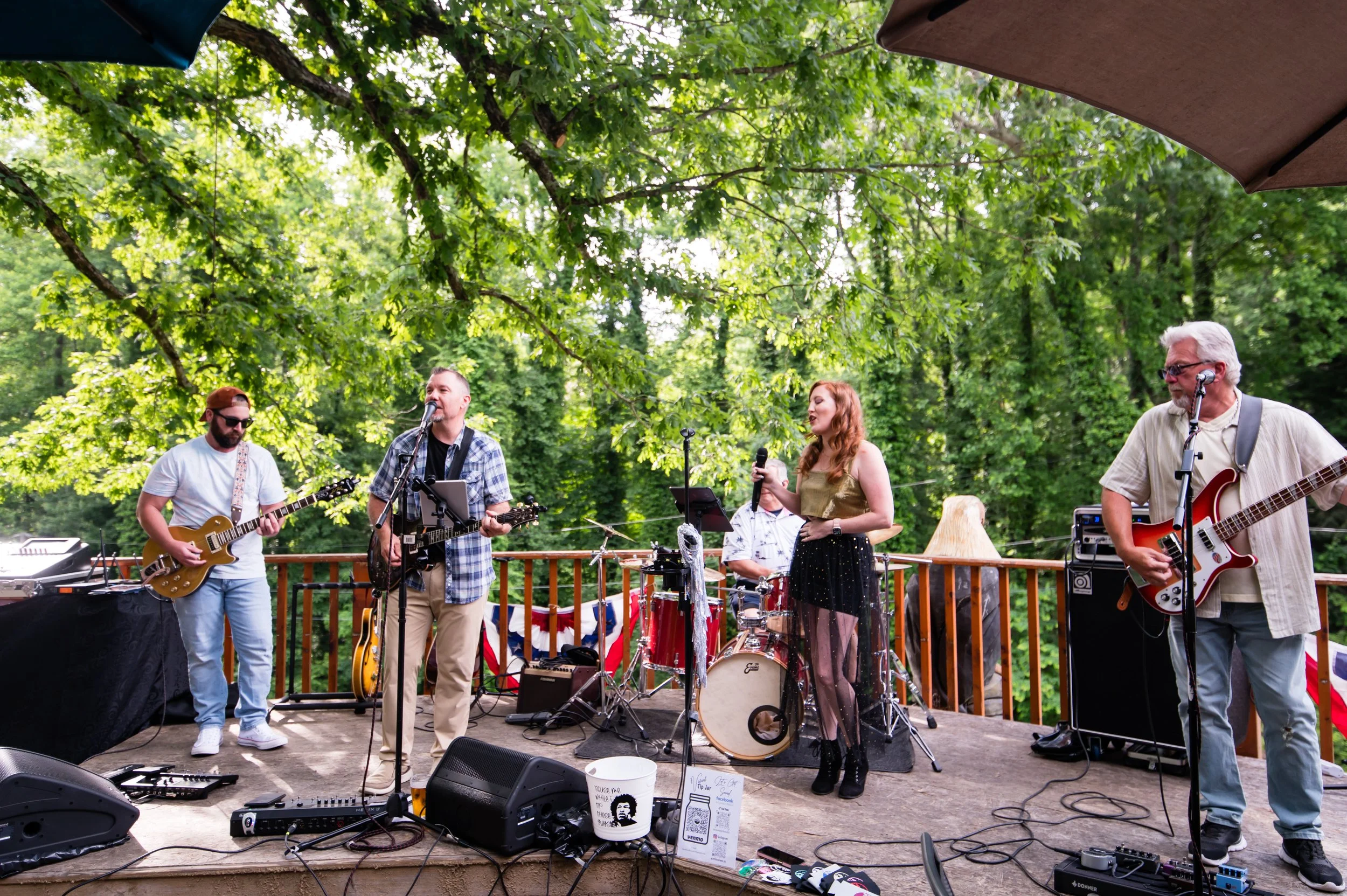 A band performing live on an outdoor wooden stage surrounded by lush green trees. The band includes a guitarist, a vocalist, a drummer, and a bassist, with various musical equipment and microphones.