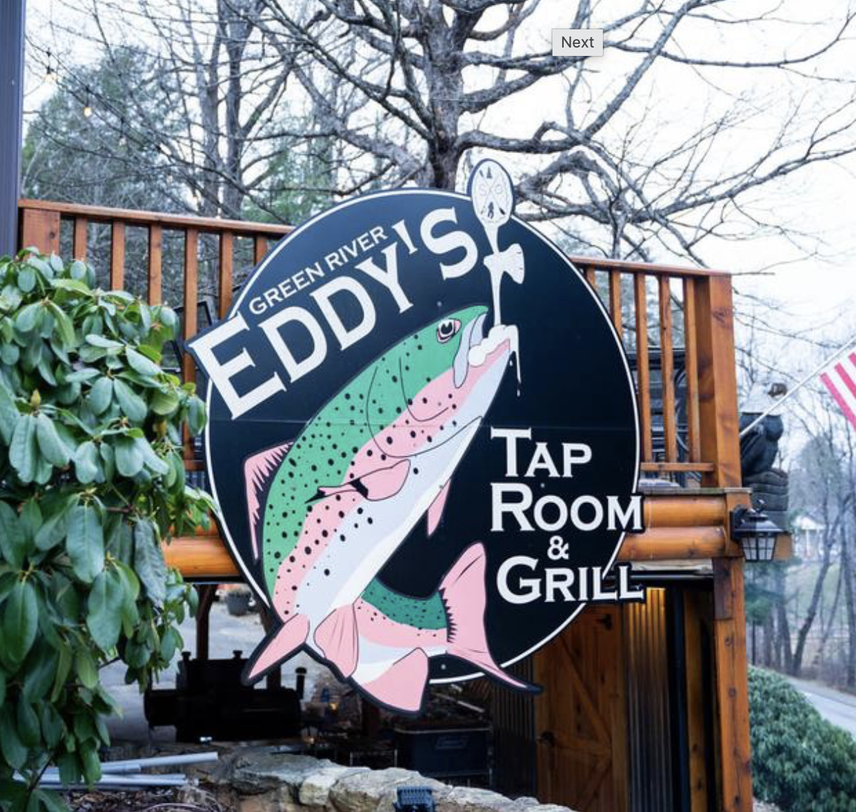 A circular sign for Eddie's Tap Room & Grill featuring a colorful illustration of a fish, mounted on an outdoor wooden deck. There are trees and greenery in the background.