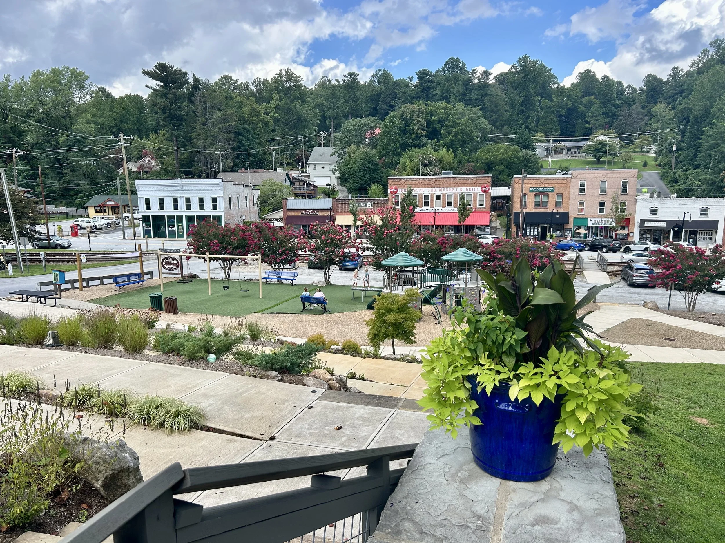 A view of a small town with colorful storefronts and buildings, surrounded by trees and hills under a partly cloudy sky. In the foreground, there is a garden with flowering plants, a blue potted plant, benches, and a play area with swings.