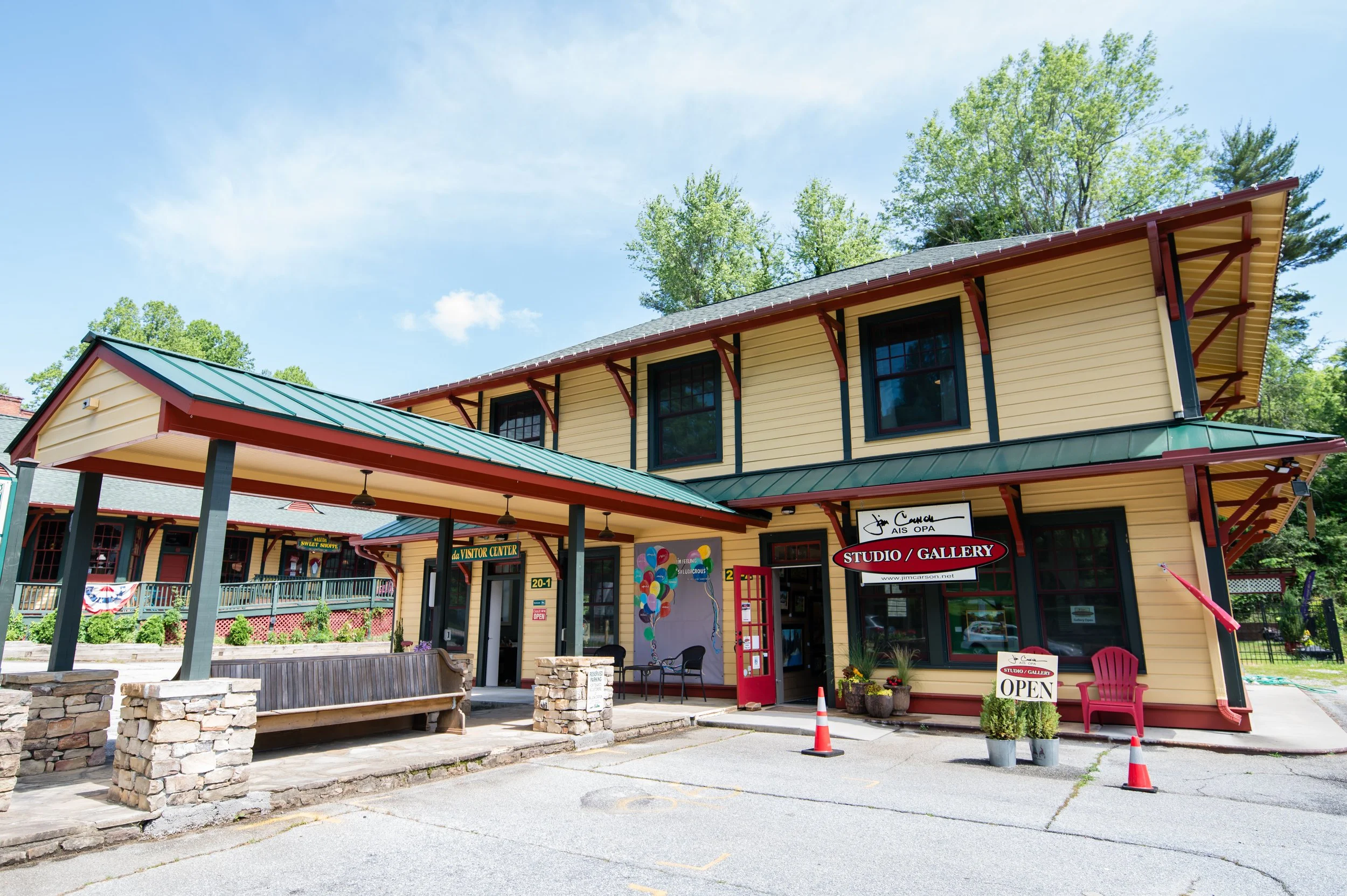 A two-story, yellow building with a green roof, housing a studio and gallery, with signs indicating it is open. In front, there is a patio with chairs, a small garden, and decorative stonework. The sky is clear and blue with some clouds, and there ar