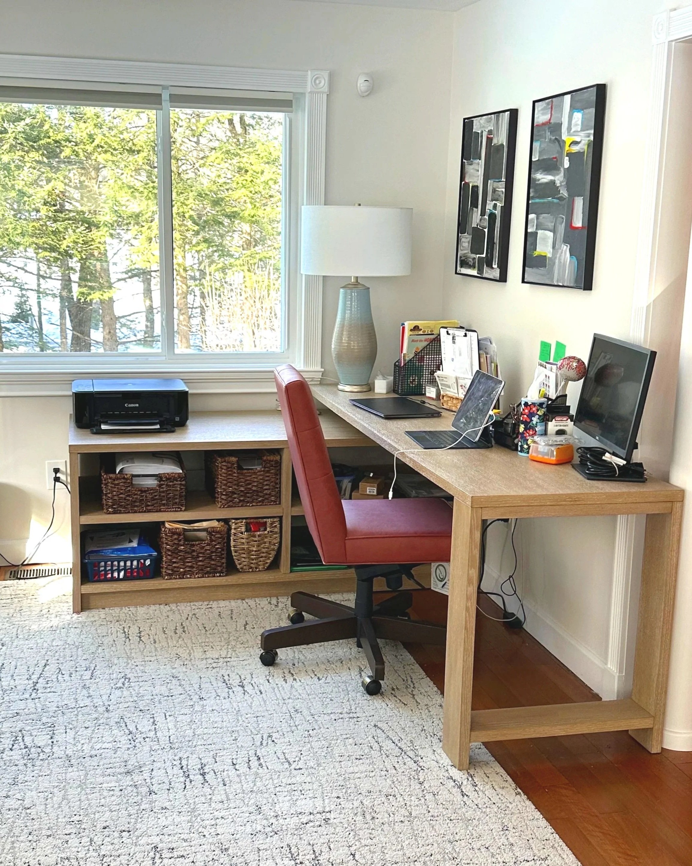 Home office with a wooden desk near a window, a red office chair, a printer on a side table, a table lamp, a desktop monitor, a laptop, and various office supplies, with framed pictures on the wall and a view of trees outside.