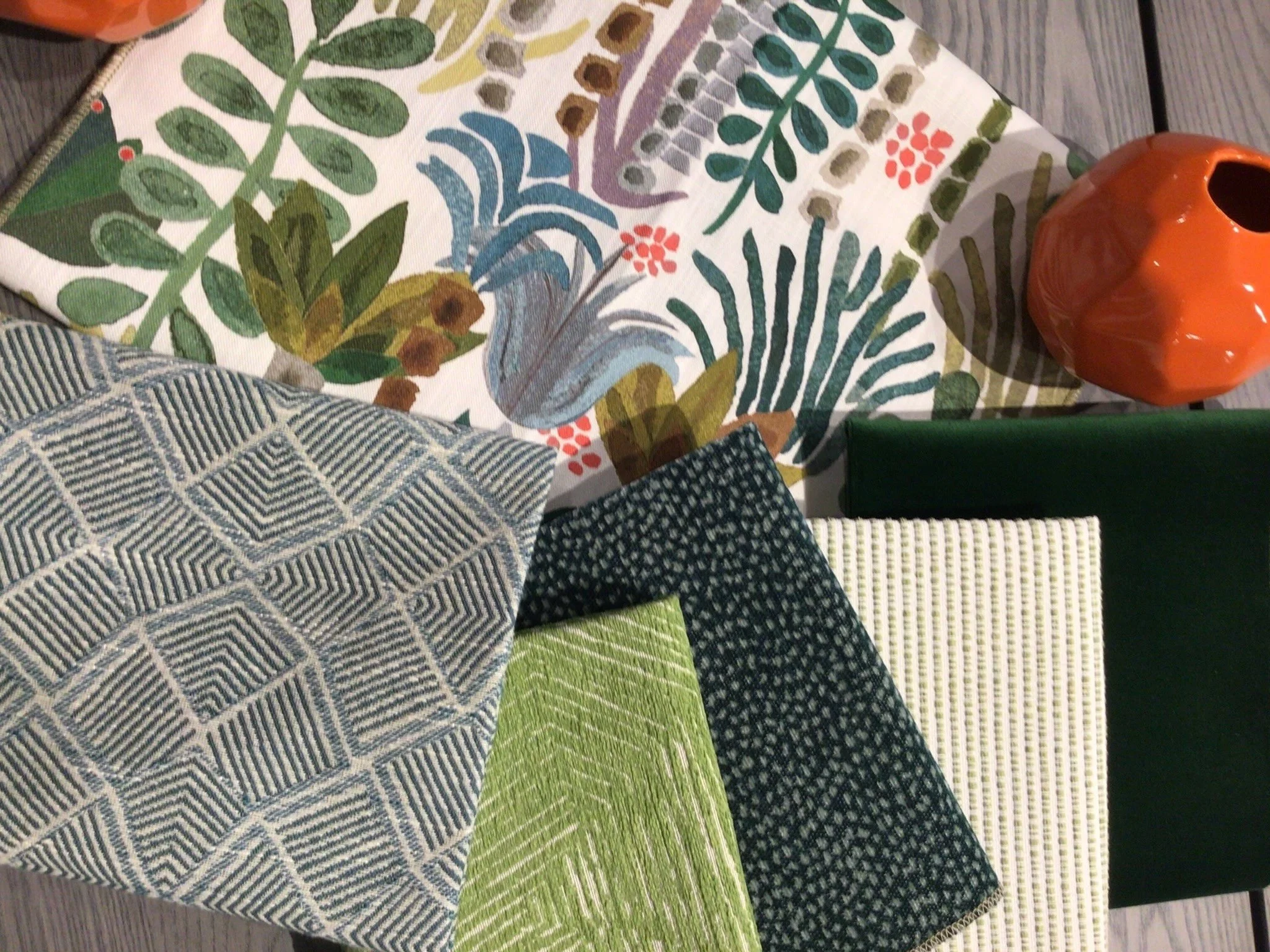 Assorted fabric swatches with various patterns and textures, including floral, geometric, and herringbone, arranged on a wooden surface, with a small orange decorative vase visible on the right side.