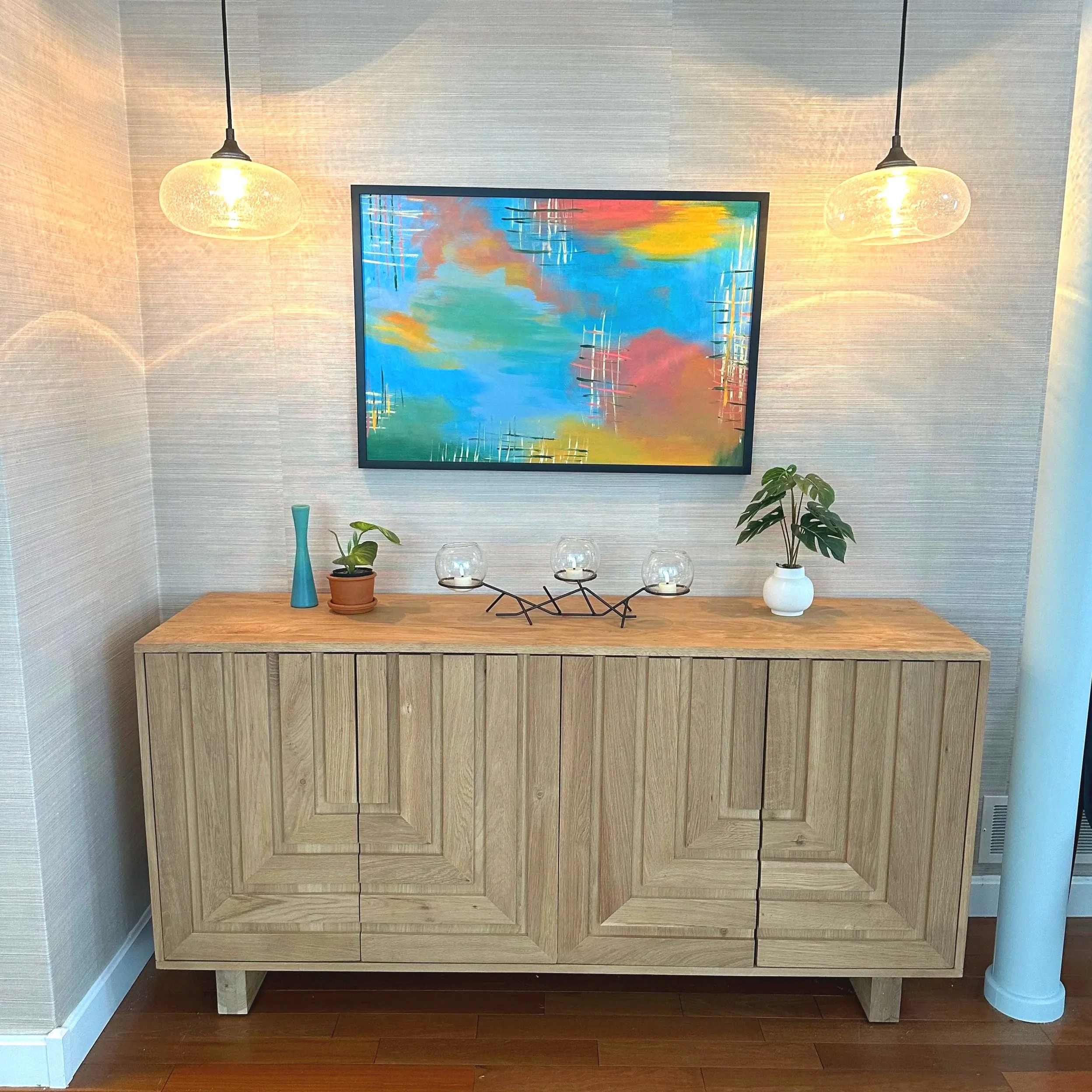 A wooden sideboard with two potted plants, glass candle holders, and a decorative metal piece on top, set against a textured gray wall with a colorful abstract painting and two hanging glass pendant lights.