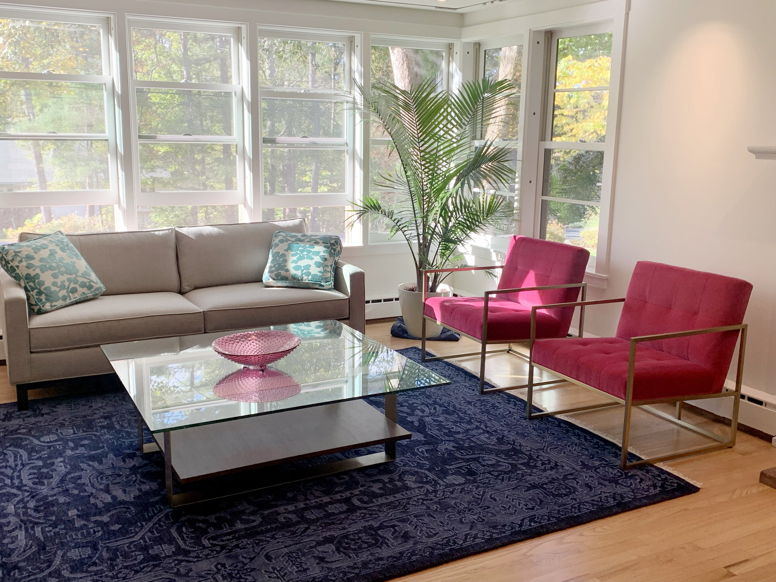 Living room with a beige sofa, two pink chairs with gold frames, a glass coffee table with a pink bowl, a large potted plant, dark blue rug, and windows showing trees outside.