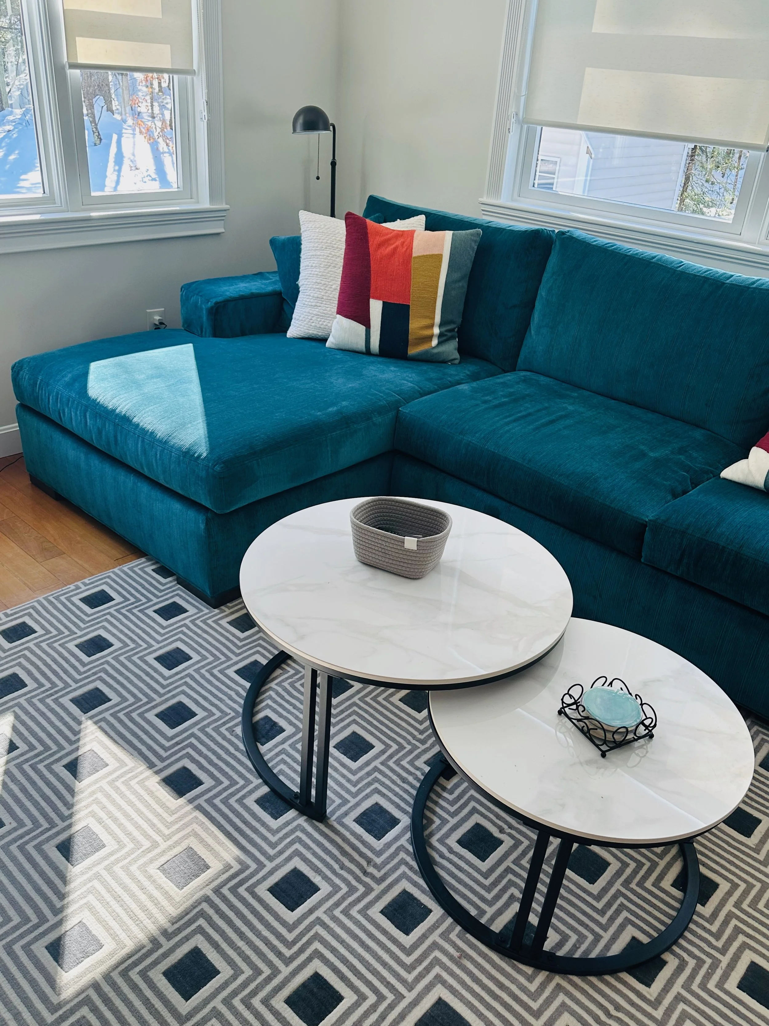 Living room with a teal sectional sofa, two white marble top coffee tables, gray geometric rug, decorative pillows, and windows with white blinds.