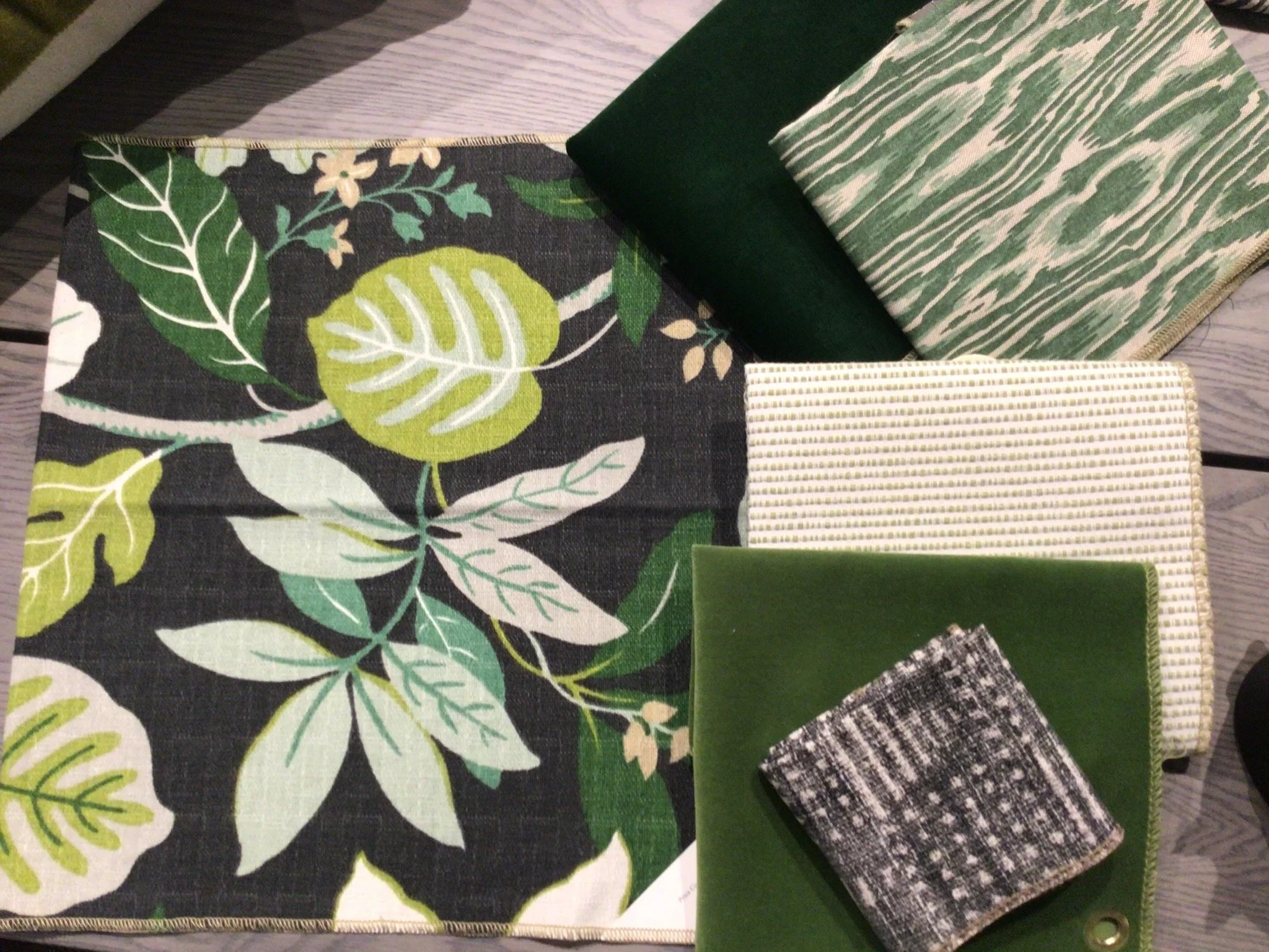 Various fabric swatches with leafy and nature-inspired patterns in different shades of green, black, white, and gray, displayed on a wooden surface.