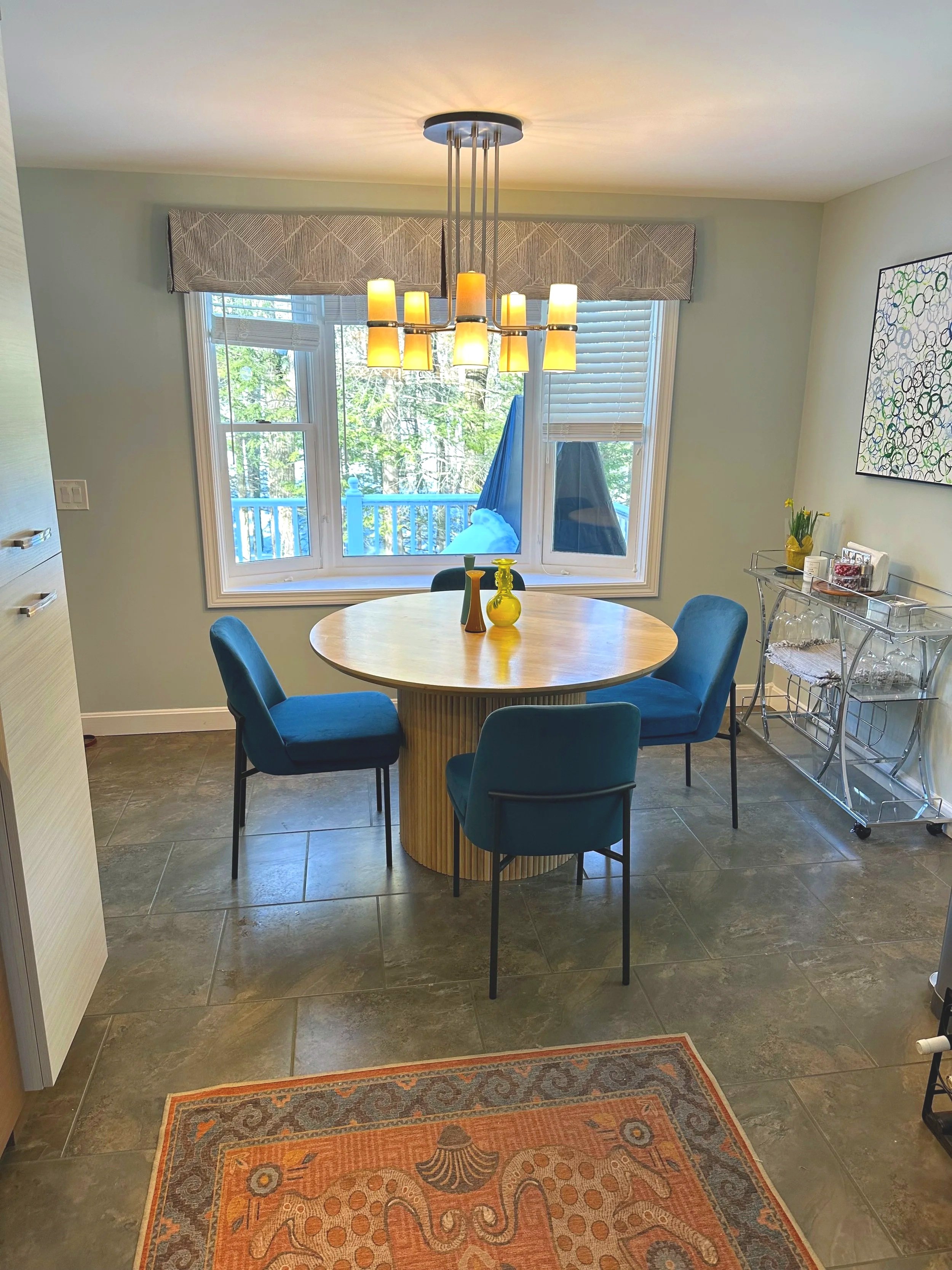 Dining room with a round wooden table surrounded by four blue upholstered chairs. A modern chandelier hangs above the table. Large window with greenery outside and a blue patio umbrella. A decorative metal cart with glassware and a wall art piece on 