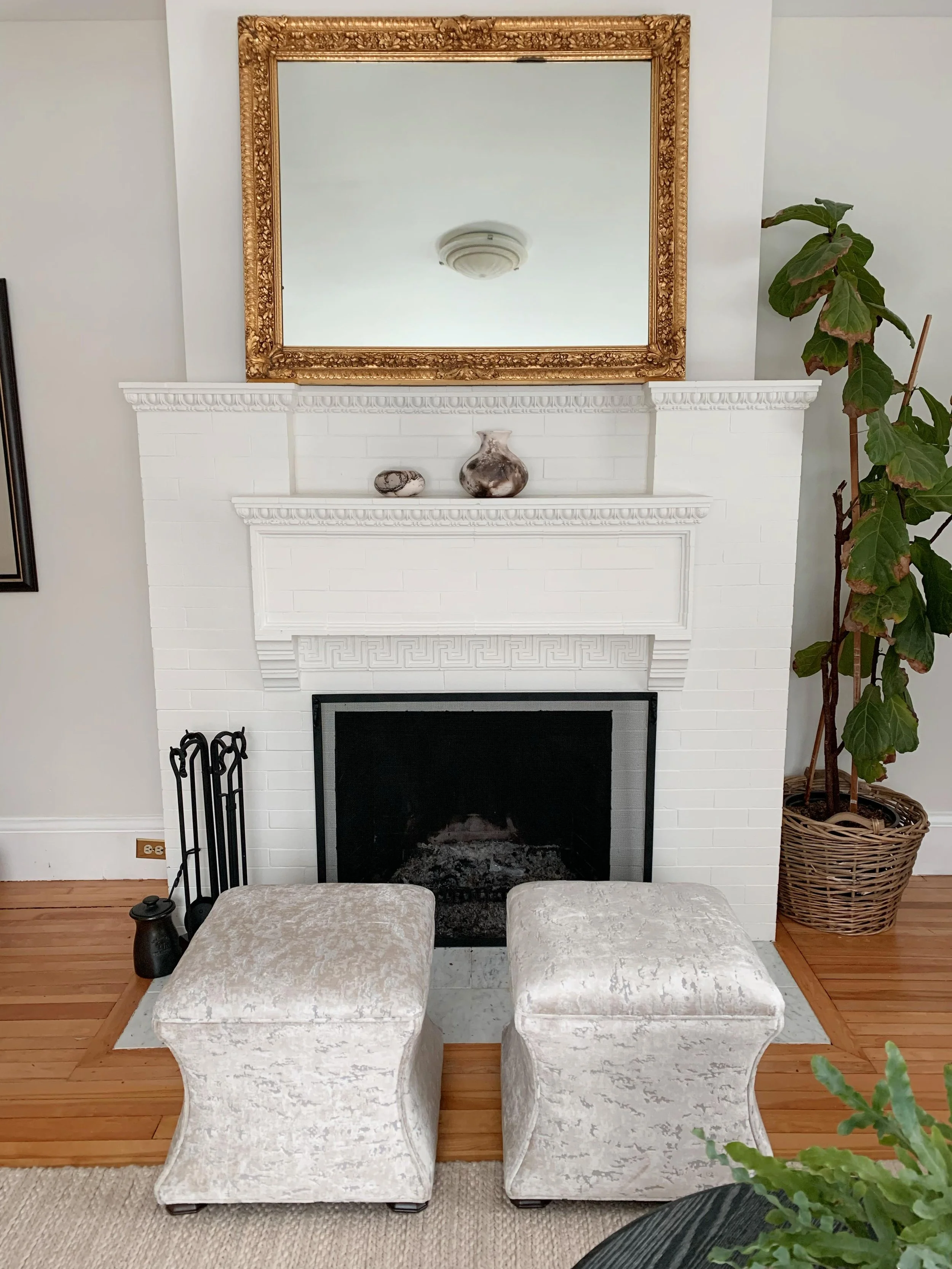 A white brick fireplace with decorative molding, a gold-framed mirror above, two small vases on the mantel, and two beige upholstered ottomans in front. There is a large potted plant to the right and a lamp and black fireplace tools on the left.