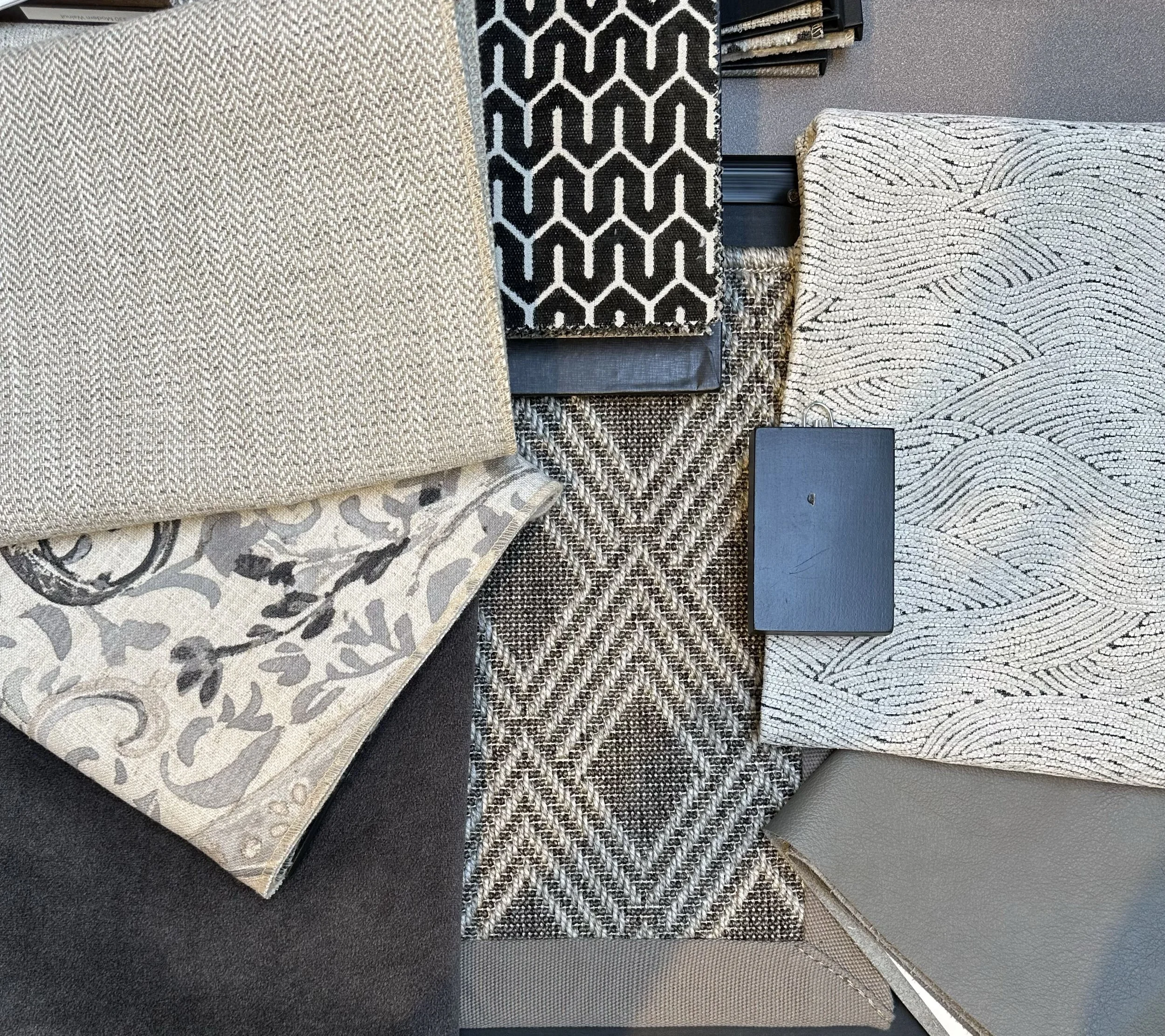 Collection of fabric samples with various patterns and textures in neutral tones, including beige, black, white, and gray, displayed on a flat surface.