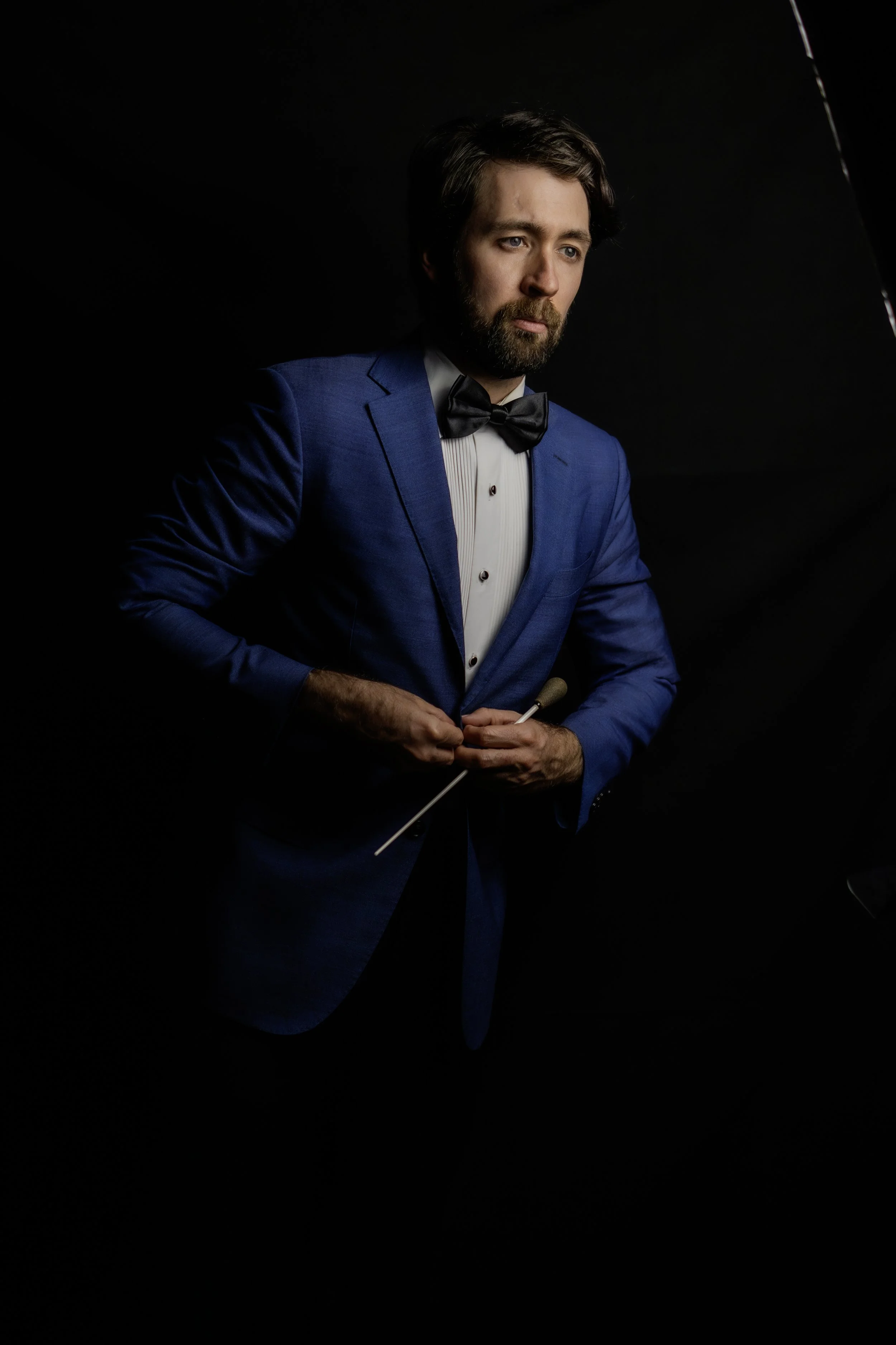 A man in a blue tuxedo and black bow tie, holding a baton, standing against a dark background.