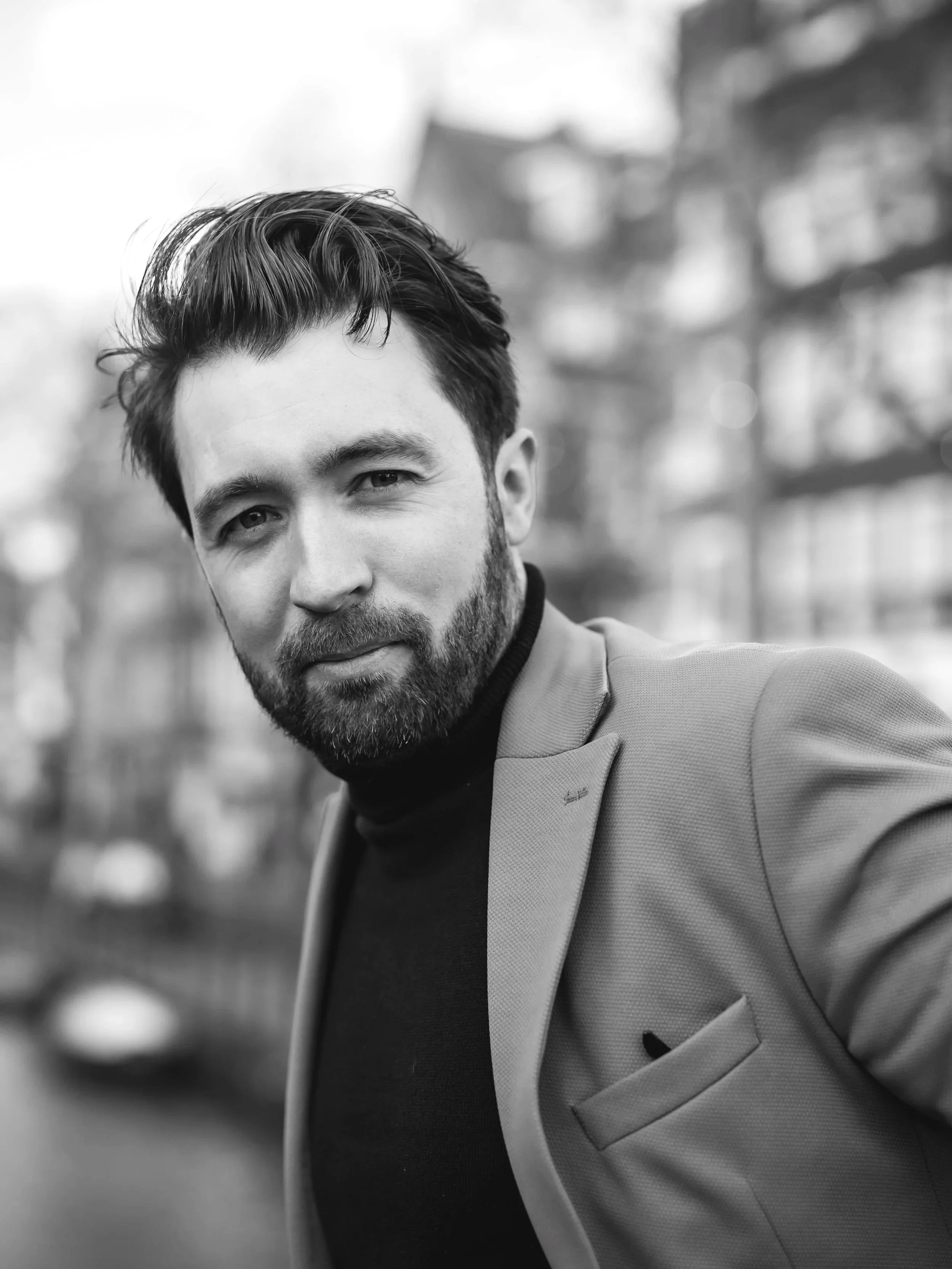 Black and white portrait of a man with a beard and styled hair, wearing a blazer over a turtleneck, outdoors with blurred Amsterdam canal buildings in background.