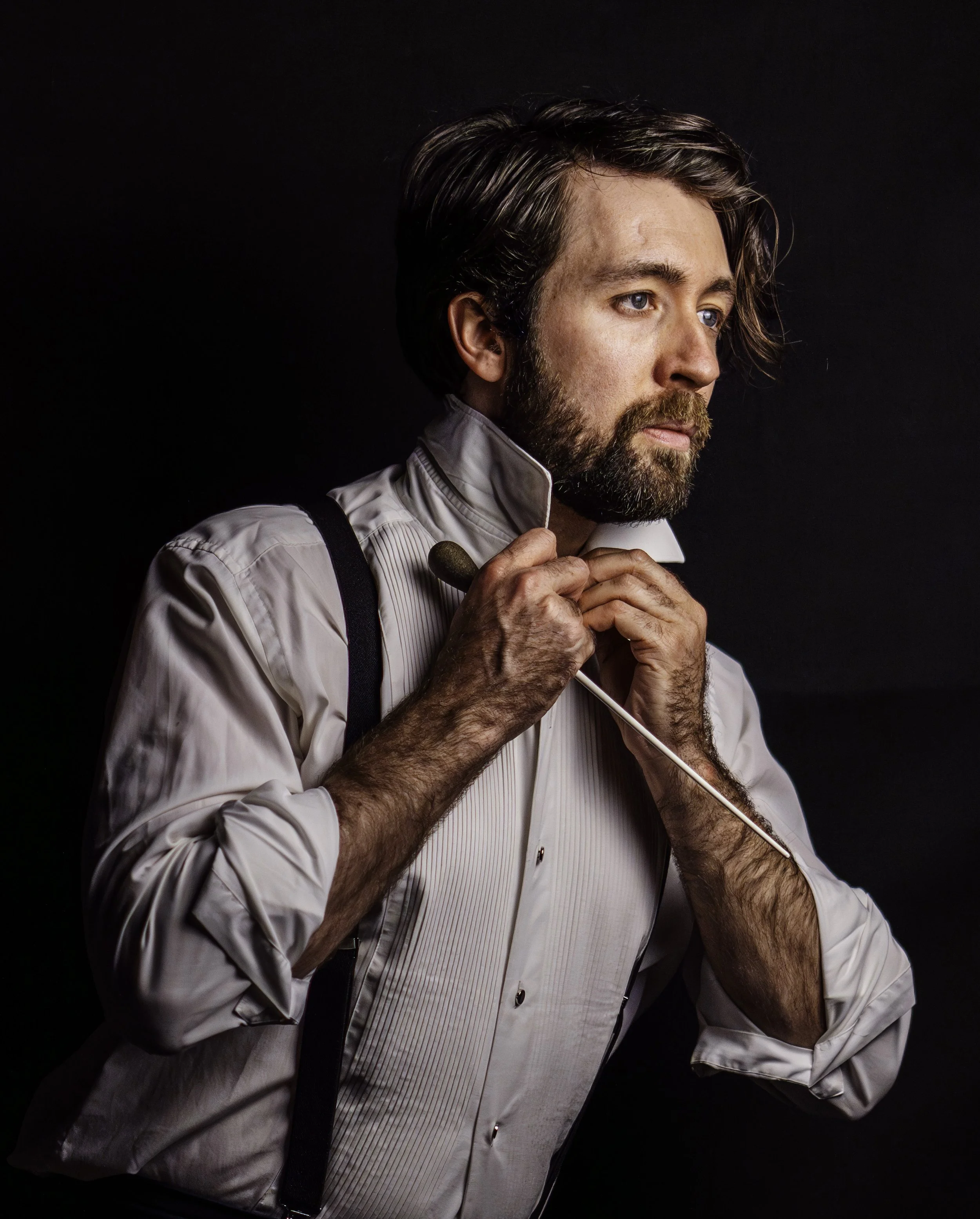 A man with dark hair and a beard adjusting his shirt collar while holding a bow tie and a conductor's baton, wearing a white dress shirt and suspenders against a dark background.