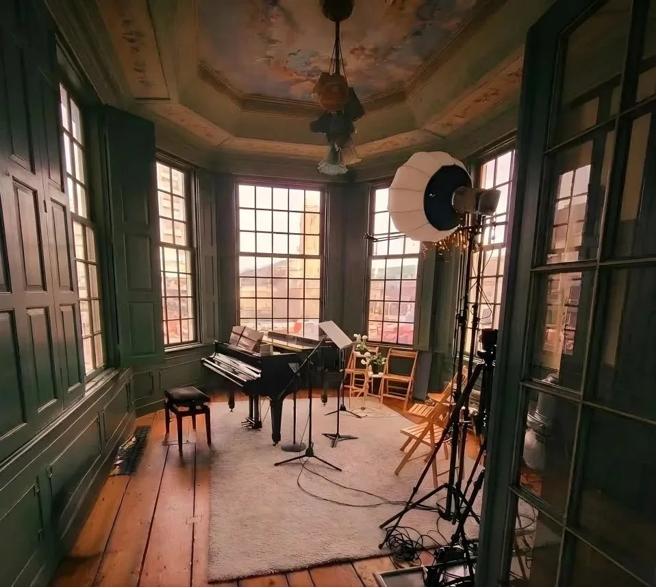 Monet Teahouse Music studio with a grand piano, music stand, microphones, chairs, and videography equipment in a room with large windows and wooden floors.