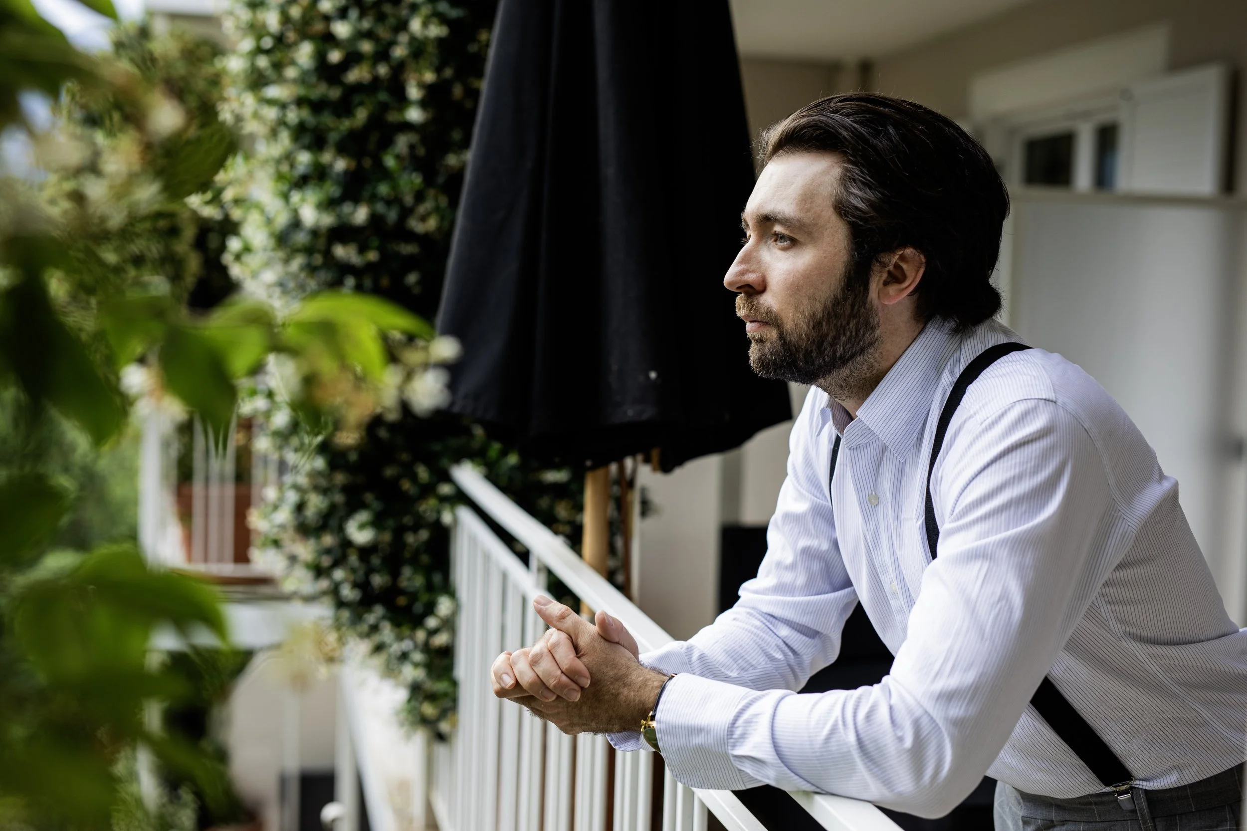 A man with dark hair and a beard, dressed in a white shirt with suspenders, leans on a balcony railing, gazing thoughtfully into the distance.