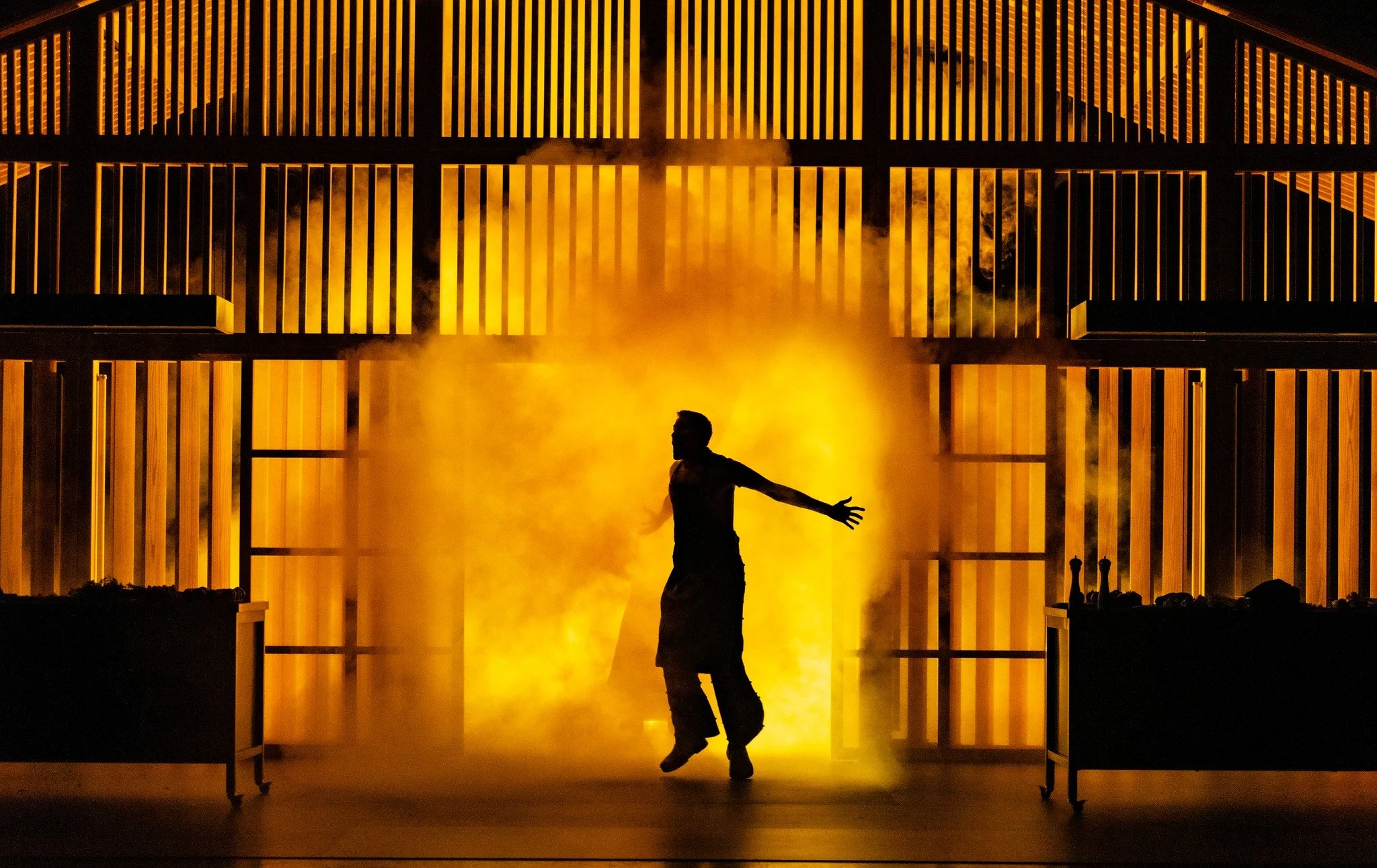 A person standing with arms outstretched in front of an intense orange flame and smoke, with a large metallic, slatted structure in the background.
