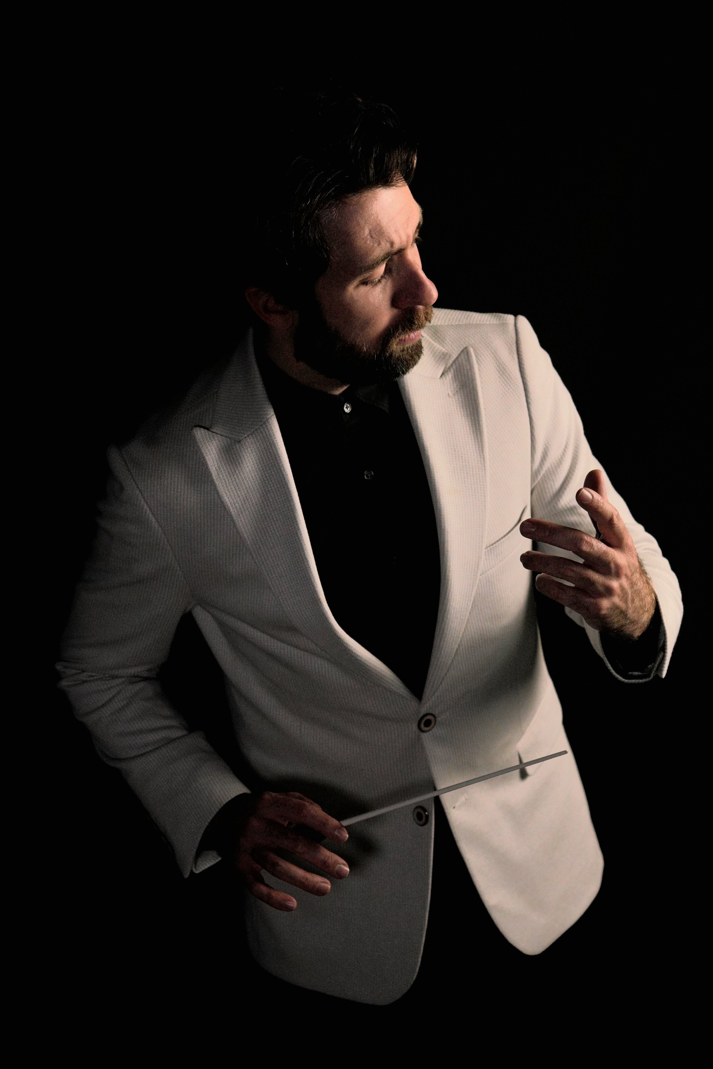A man with a beard and dark hair, wearing a white blazer and black shirt, is conducting with a baton against a black background.