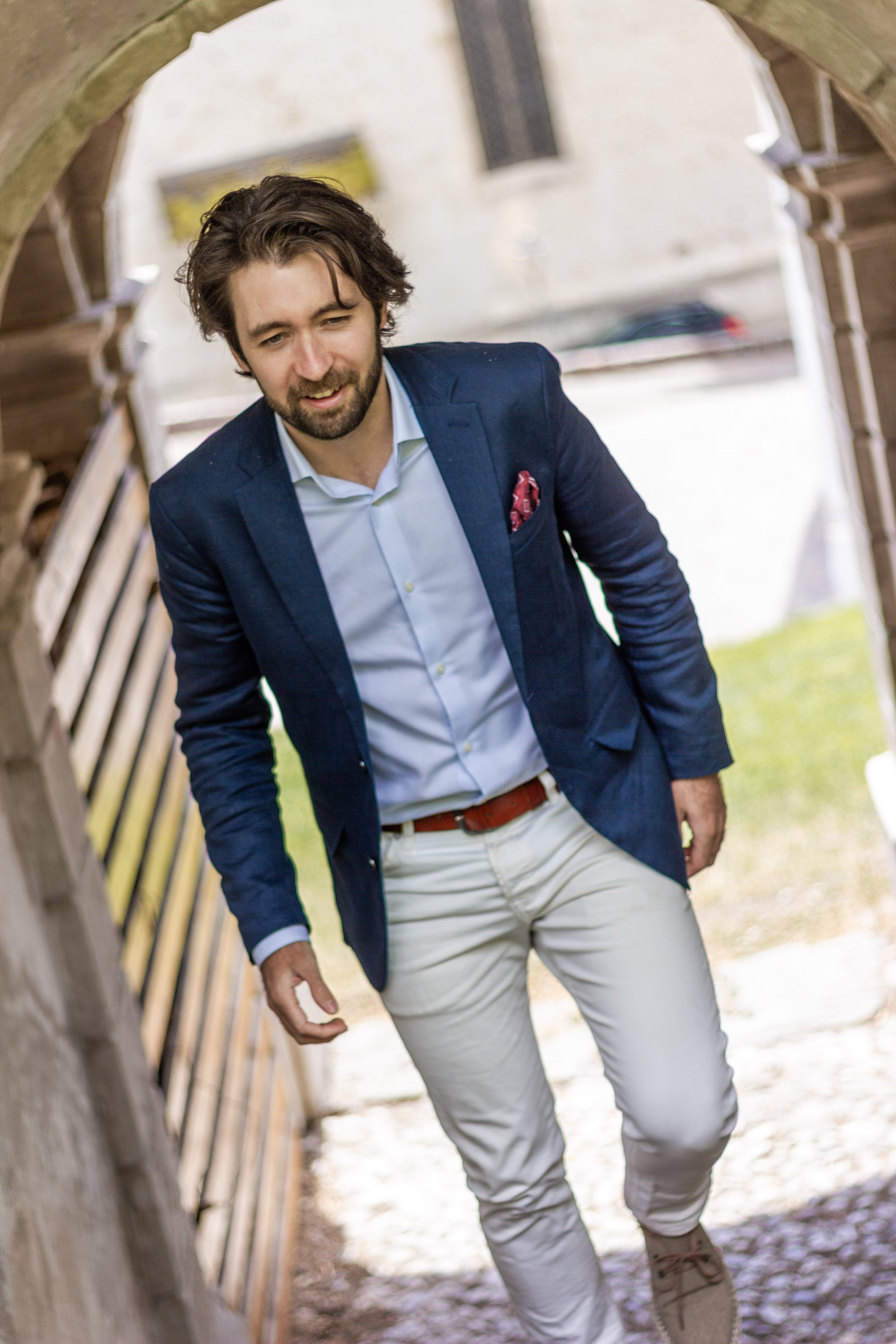 A man in a blue blazer, white shirt, and white pants walking through a stone archway outdoors, smiling.