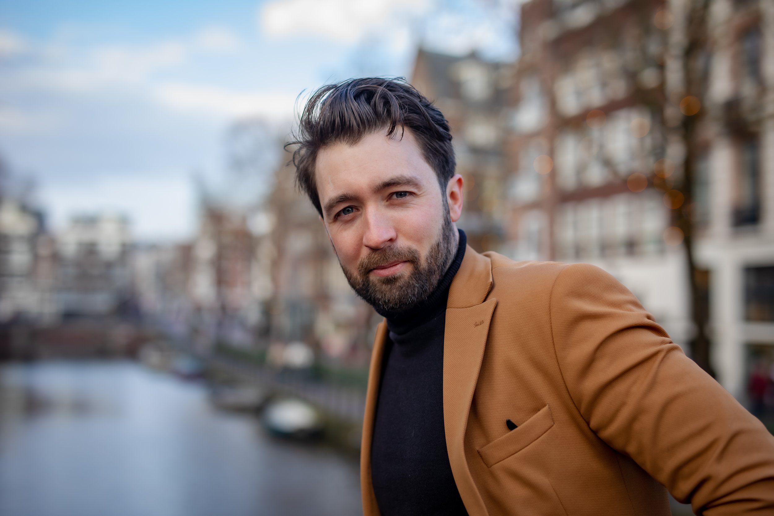 A man with dark hair and a beard taking a selfie outdoors near a canal, wearing a tan blazer and black turtleneck, with city buildings and trees in the background.