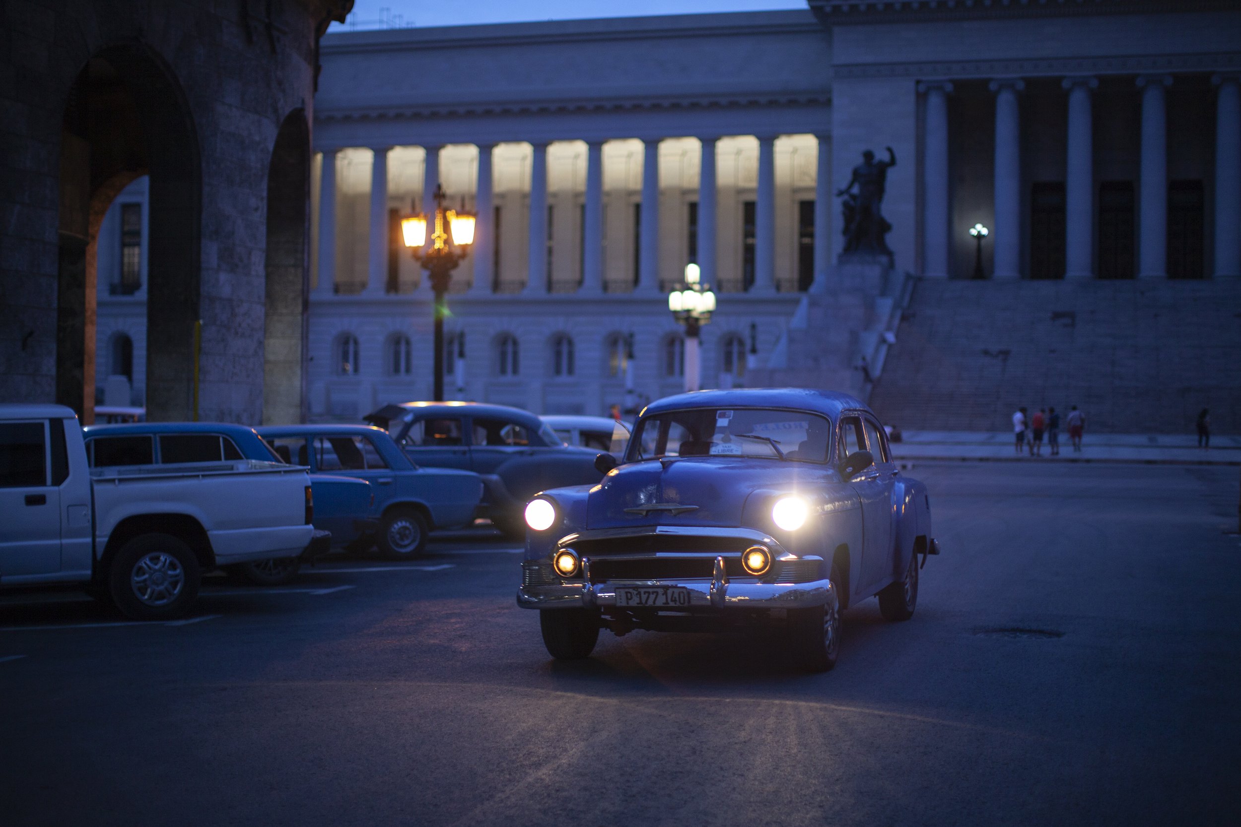 Habana nights, Cuba. Vintage car with headlights on driving through a city square at dusk, with old buildings and statues in the background.