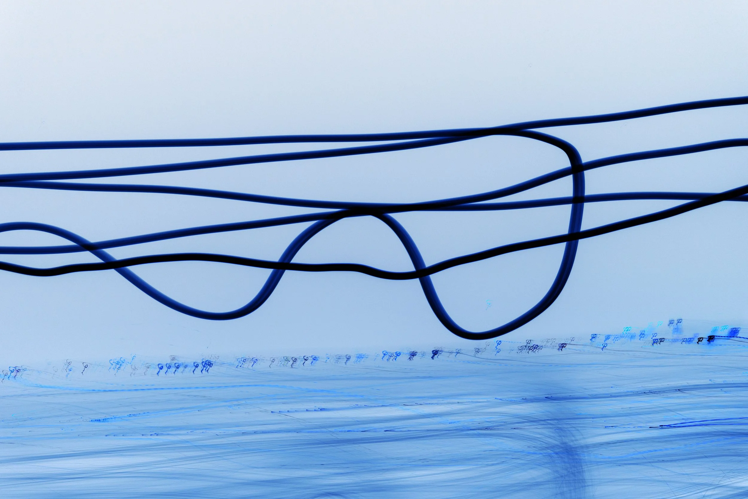 A close-up of hanging blue electrical wires against a light sky, with a blurred landscape of more wires and poles below.