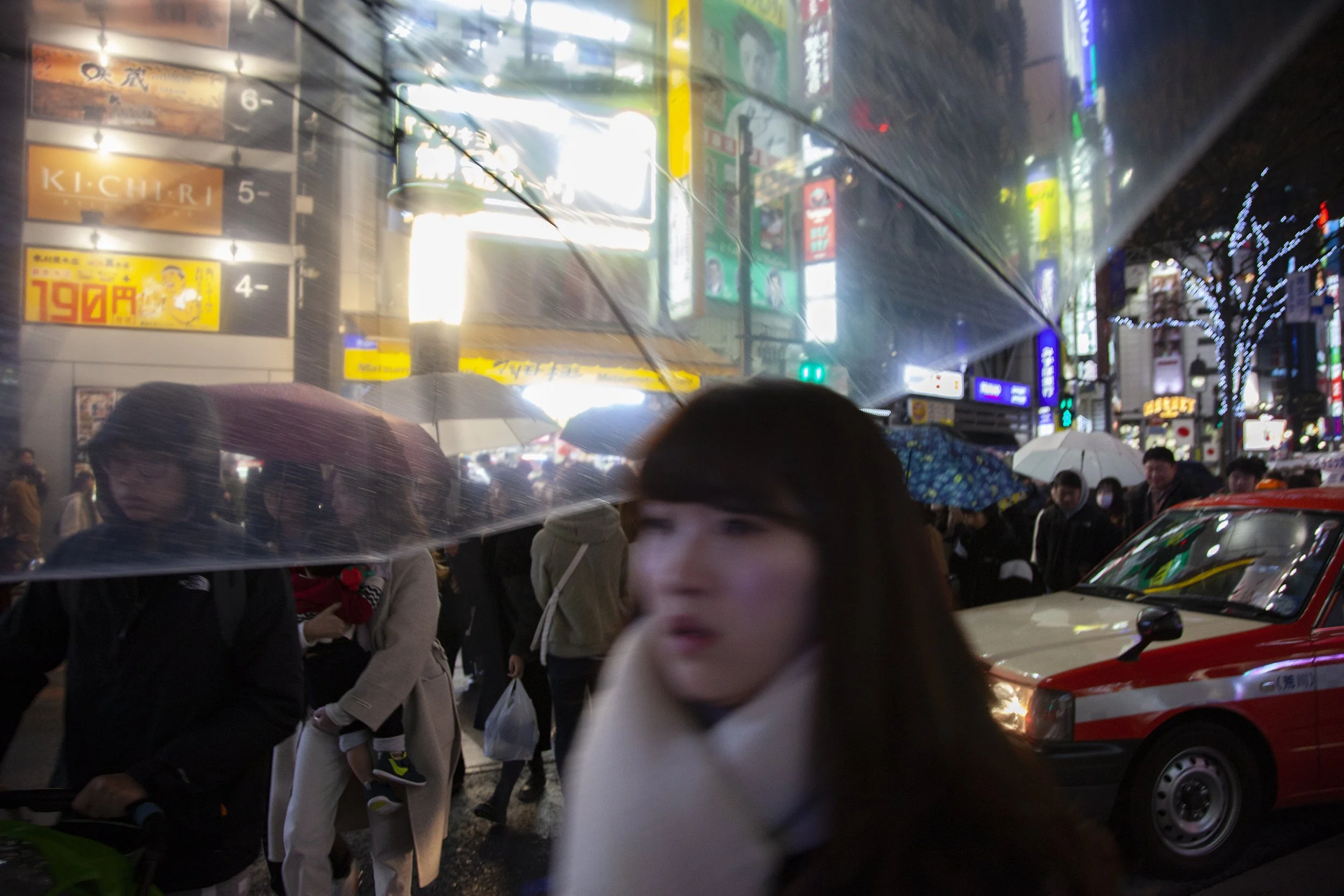 A busy city street scene at night with people holding umbrellas, flashing neon signs, and a taxi cab in the foreground.