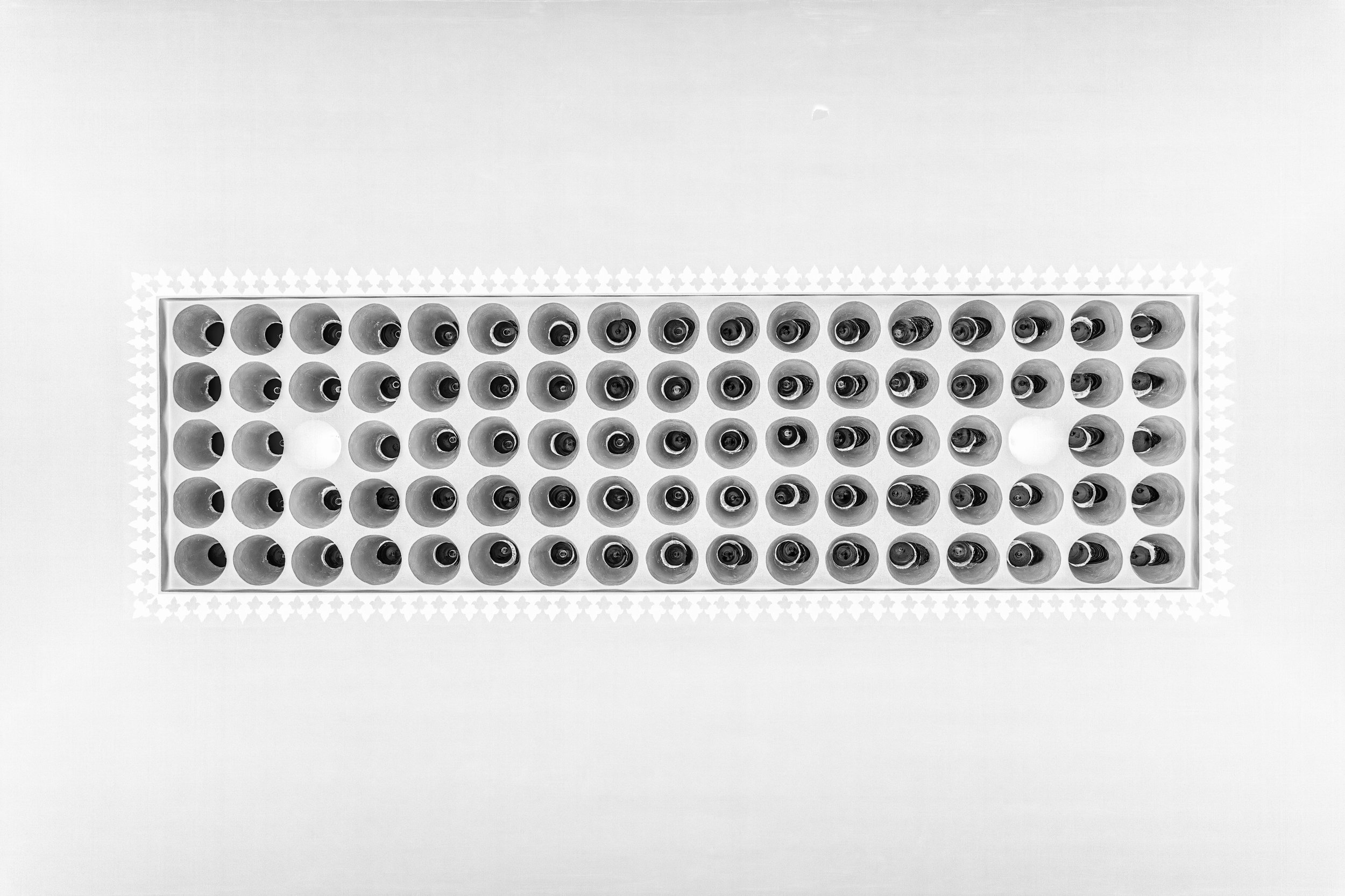 Top-down view of a rectangular container with numerous small bottles or vials arranged in neat rows, with a few bottles missing, on a white surface.