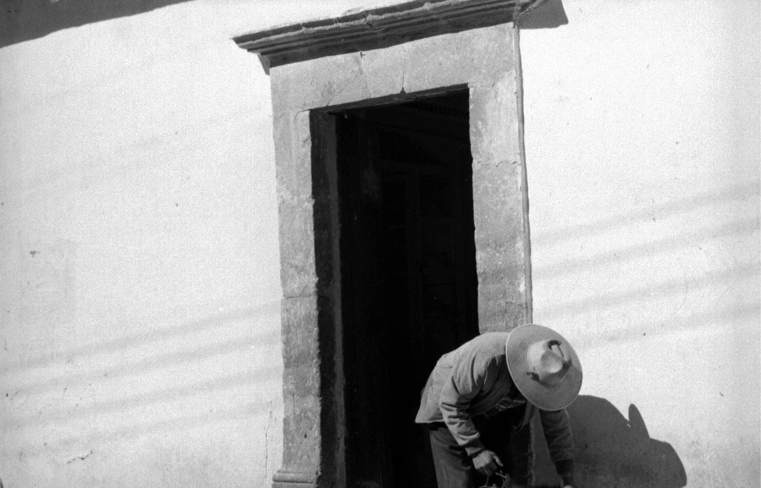 A person wearing a hat is crouched by a stone door frame on a white wall, working on the ground. Old Mexico