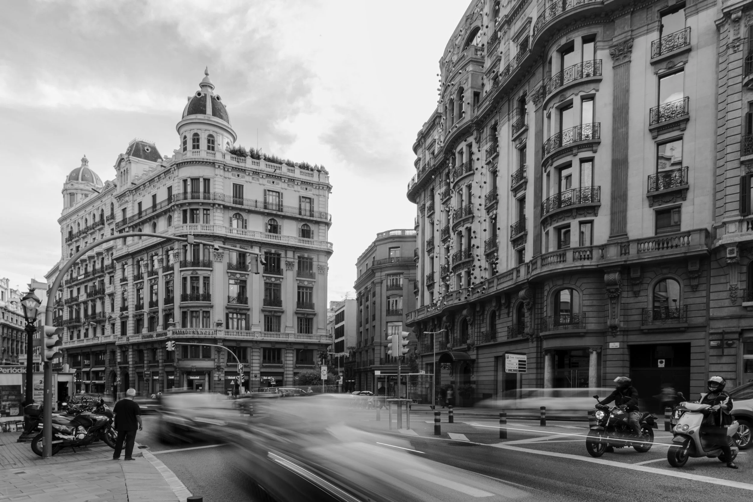 Black and white photo of a busy city street with tall historic buildings, cars and motorcycles, and a person waiting on the sidewalk.