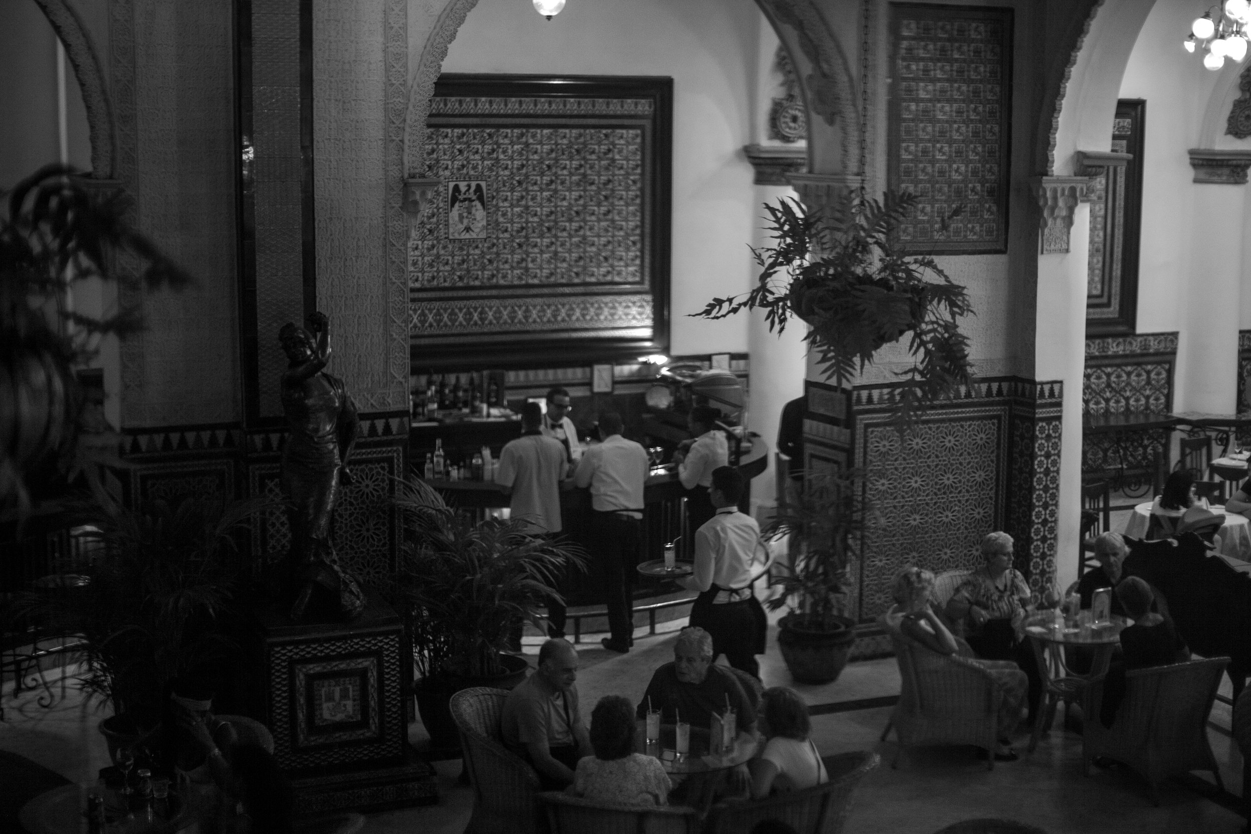 Habana nights Cuba. A black and white photo of a lively, elegant restaurant with ornate architectural details, large potted plants, and groups of people dining and socializing.