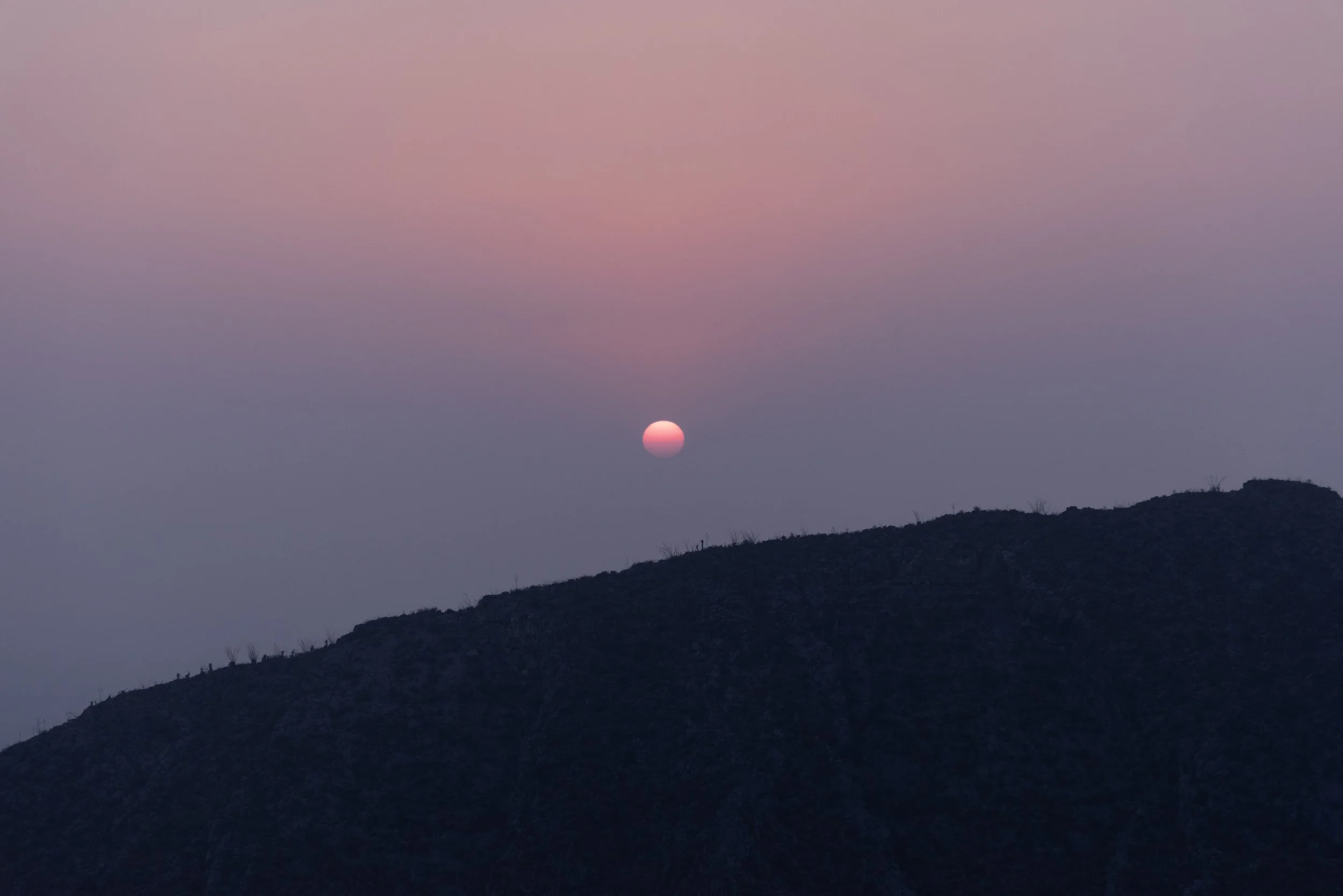 A pink sun setting or rising over a dark mountain silhouette under a pastel pink and purple sky.