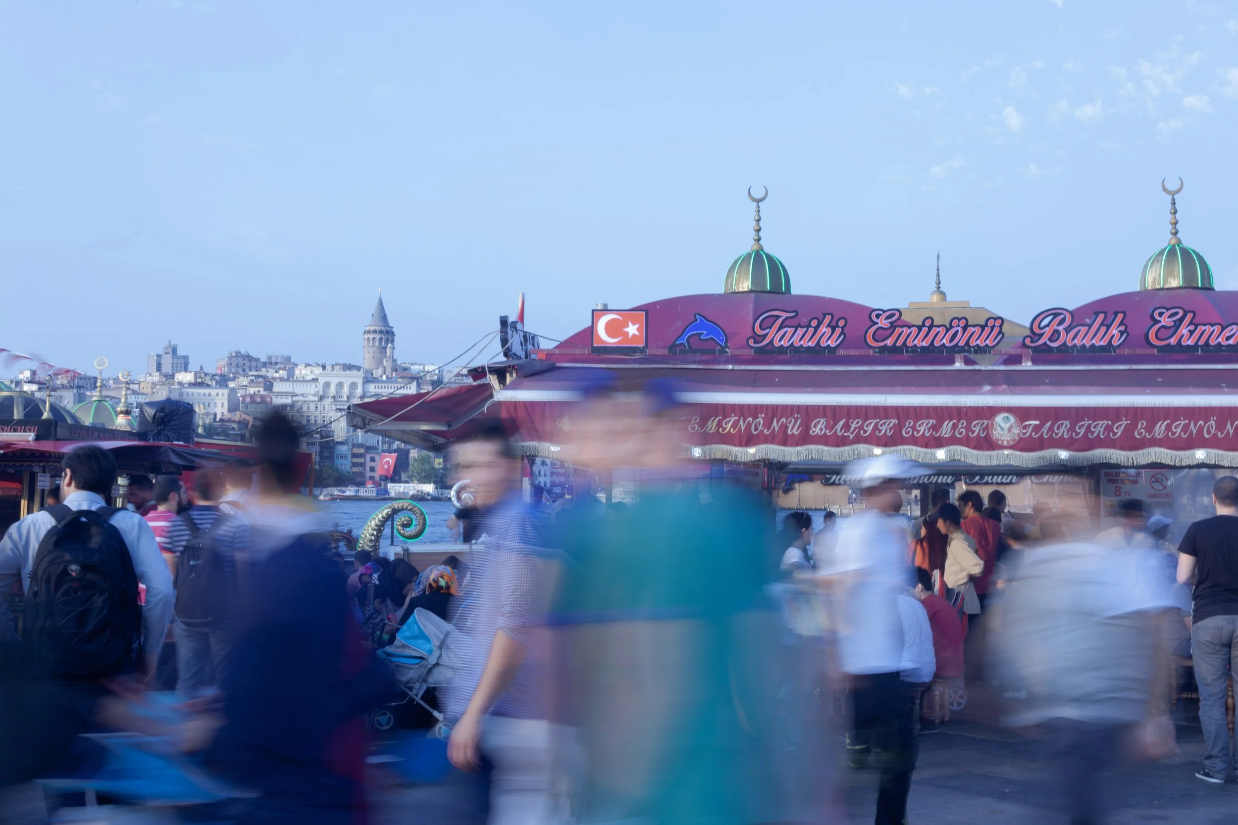 A bustling outdoor market scene with many blurred pedestrians walking amidst colorful stalls during daytime. A large red awning displays language in Turkish and symbols including a Turkish flag and a dolphin icon. The background shows a cityscape with historic buildings and a tower on a hillside under a partly cloudy sky.