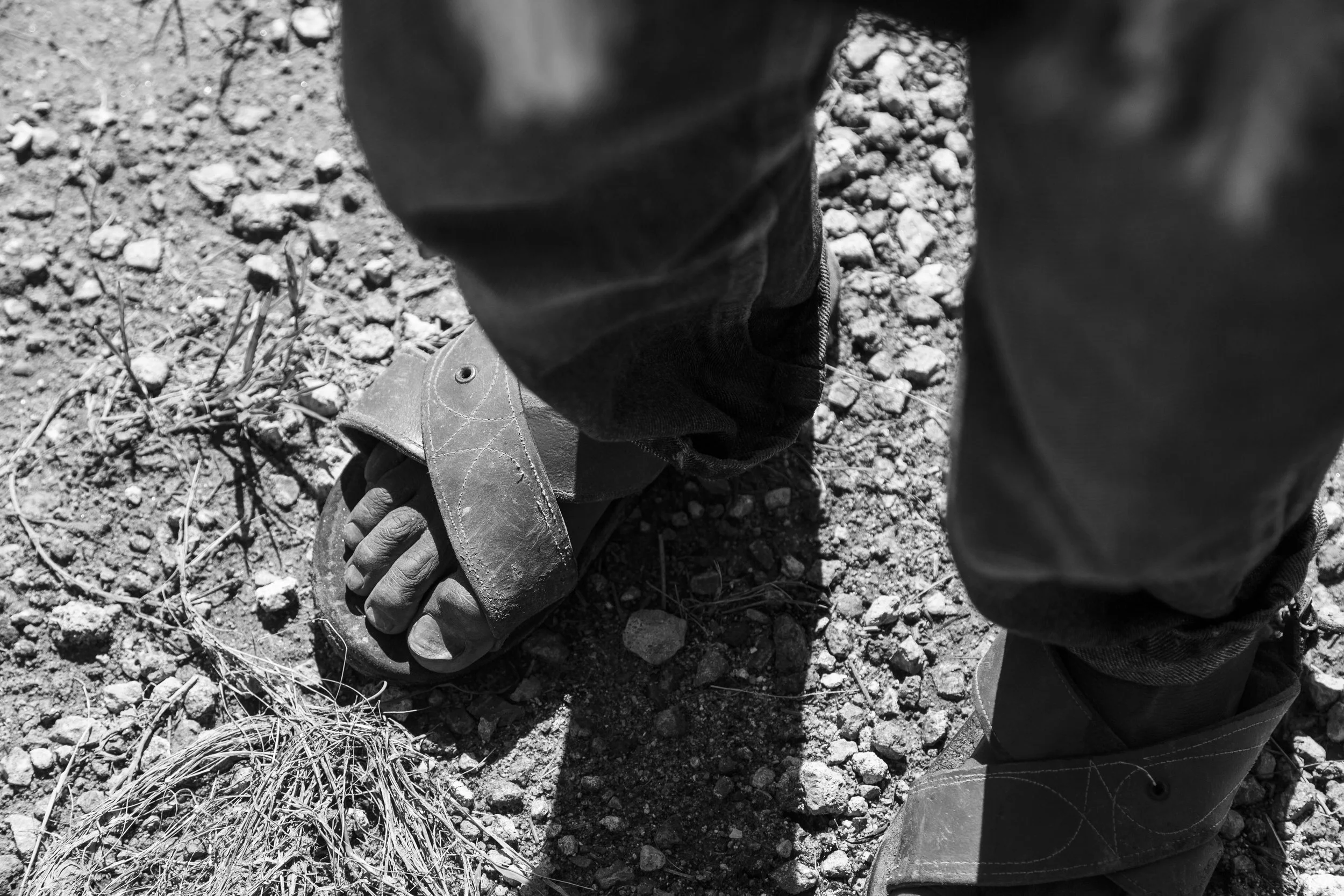 Mexico presences. Old and ancient. Close-up of a person's feet wearing sandals and jeans, standing on rocky ground with dry grass in sunlight.
