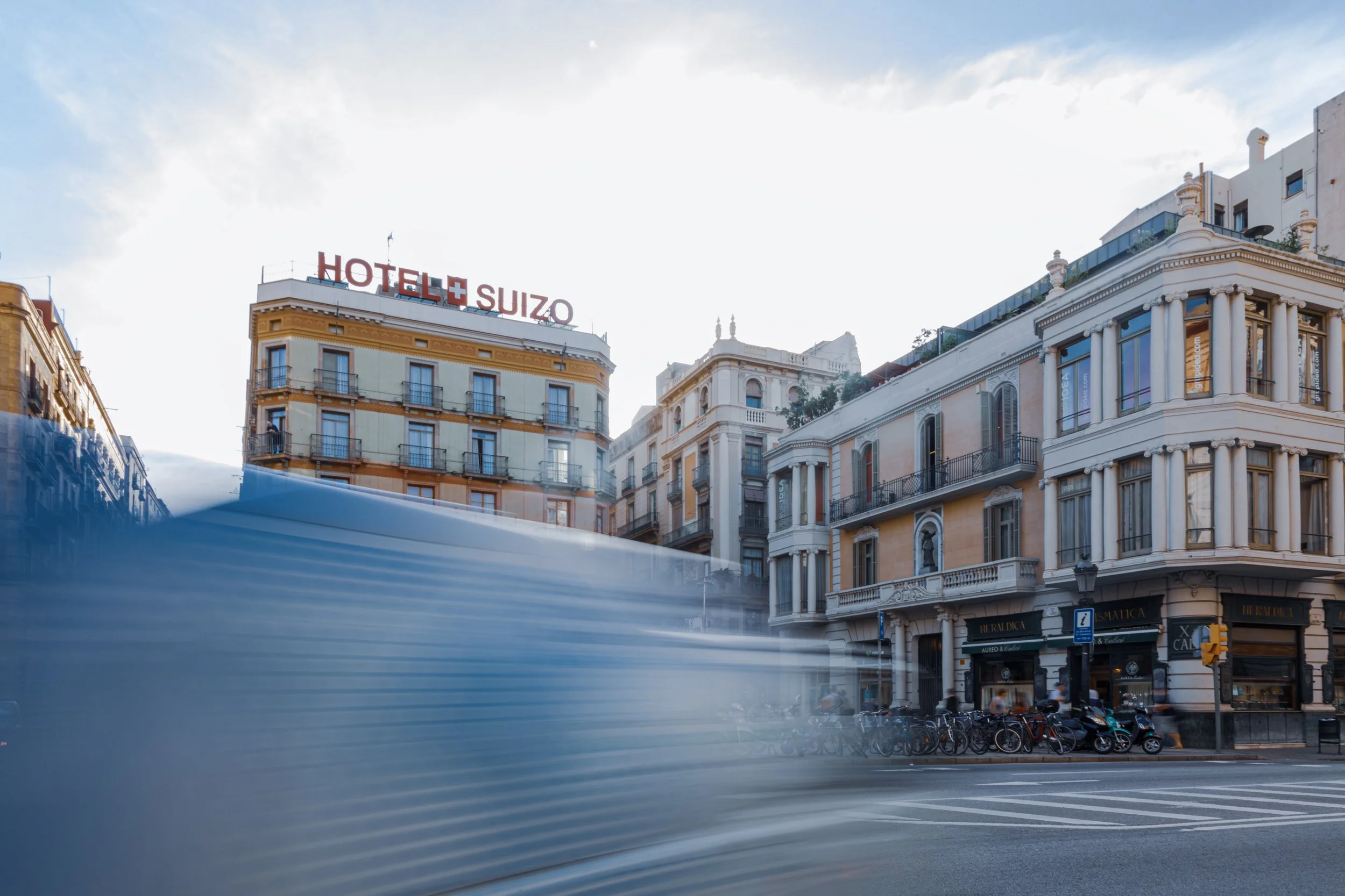 City street view with historic buildings, a hotel sign, bicycles parked, and a blurred vehicle passing by at crosswalk during daytime.
