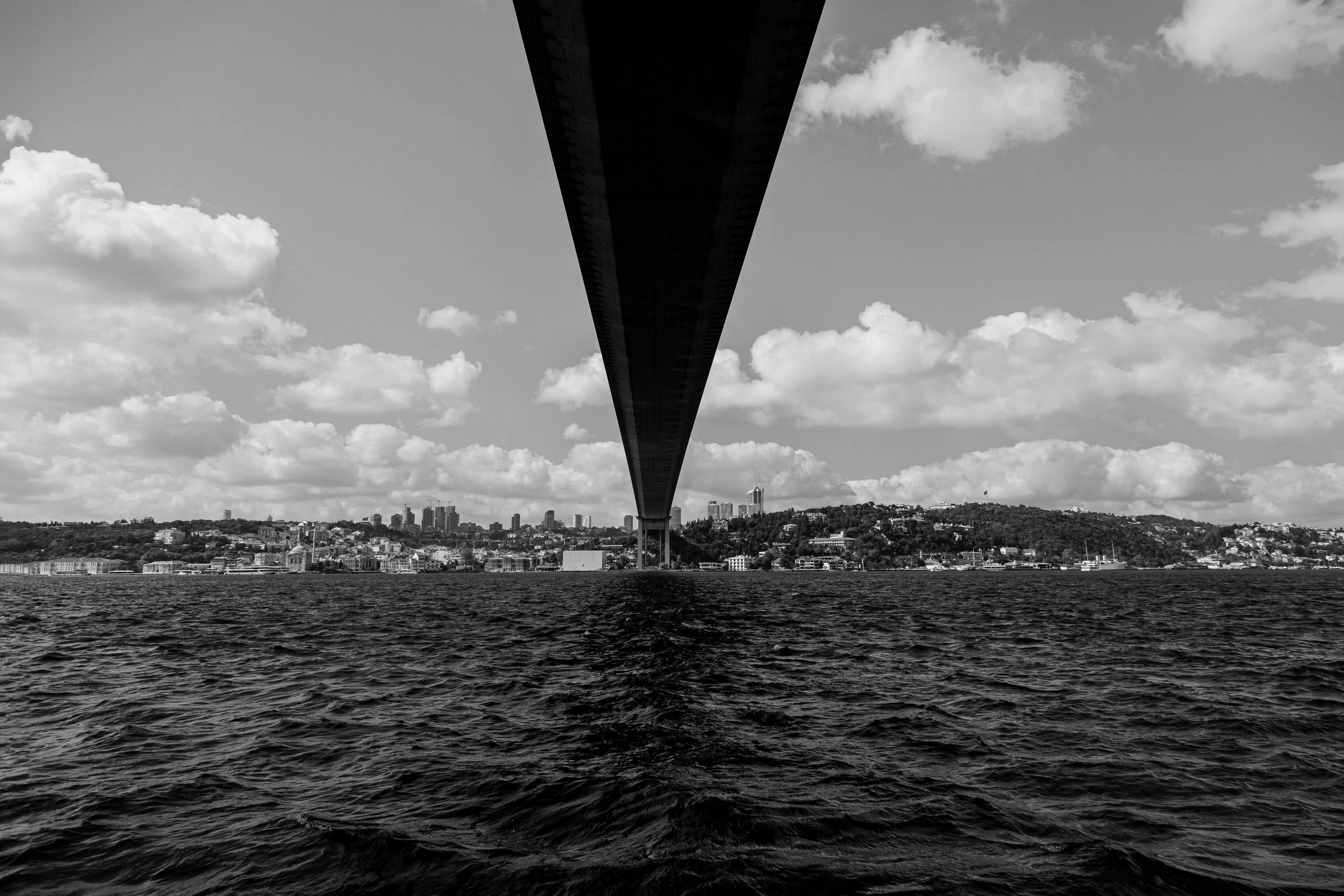 Black and white photo of a bridge viewed from below over the water with a city skyline and hills in the background.