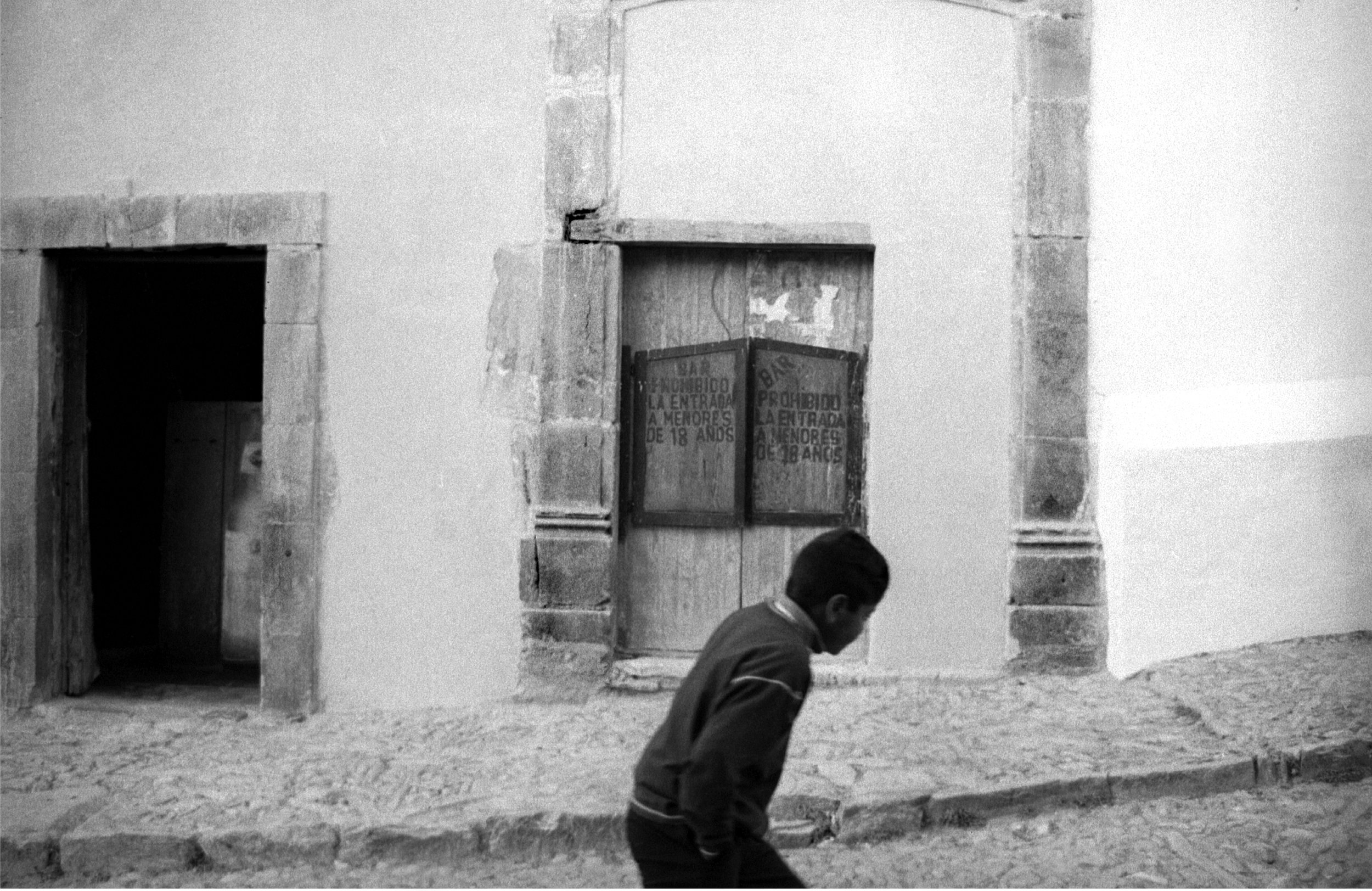 A boy walking past a building with a boarded-up window and an open doorway, in an urban setting. old Mexico