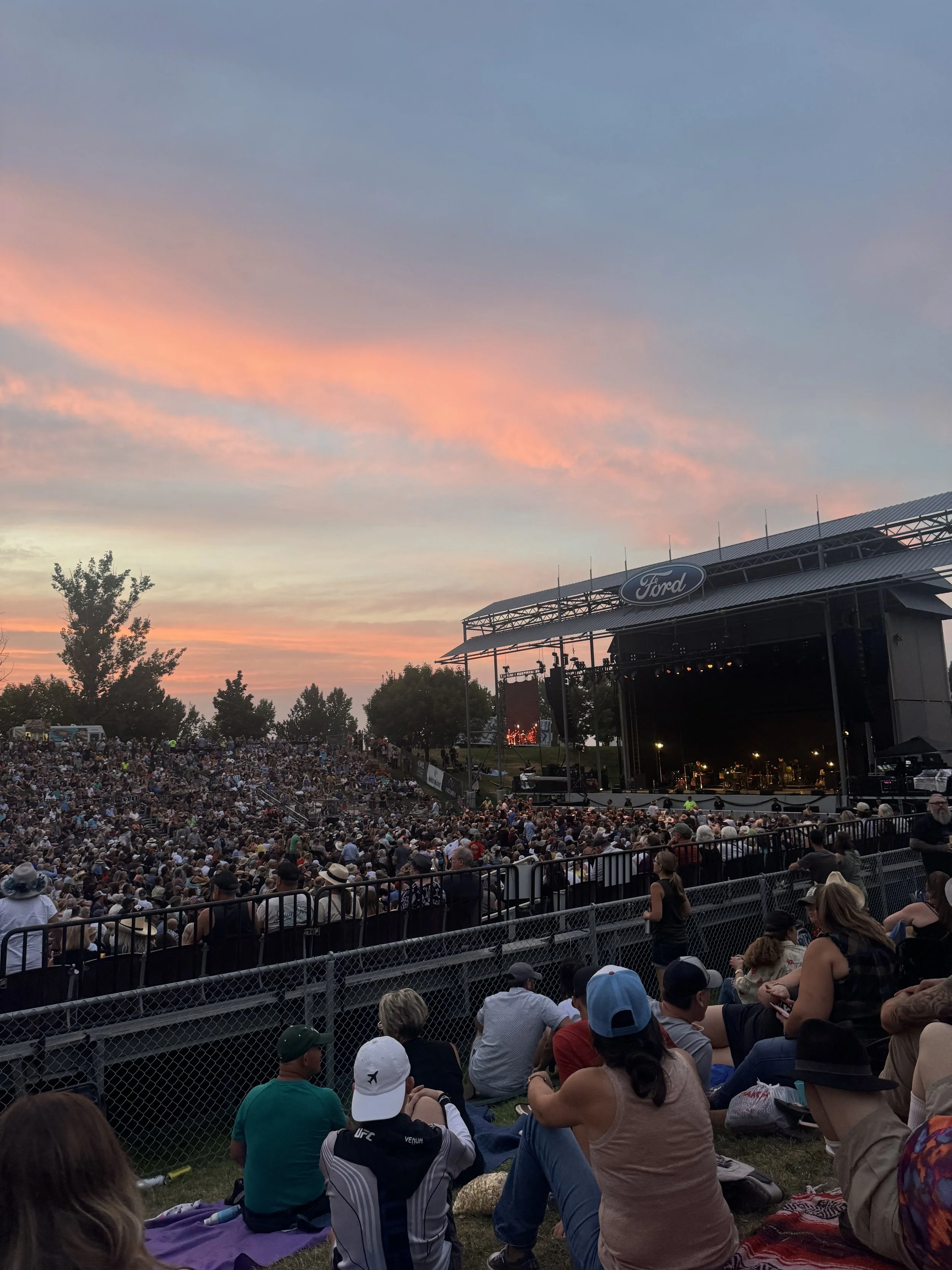 An outdoor concert at sunset with a large crowd seated in a stadium. The stage has a Ford logo and a colorful sky with pink and blue clouds.