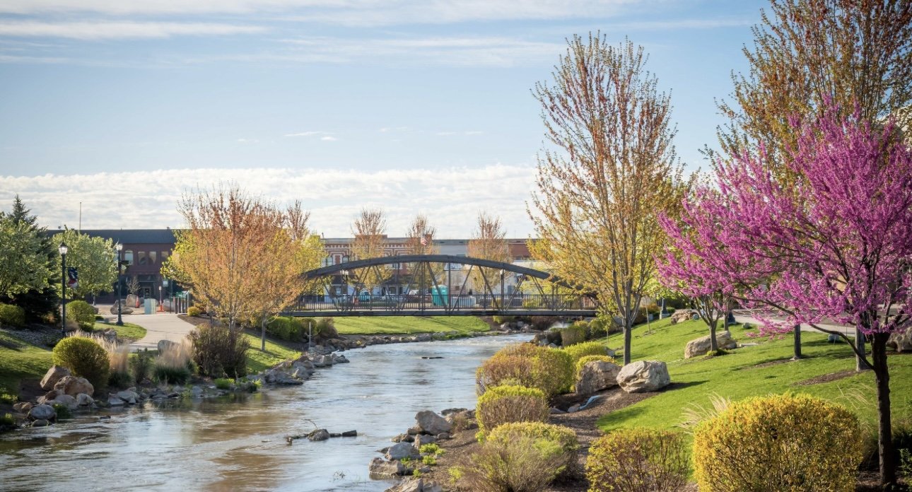 A peaceful park scene with a small river flowing through it, surrounded by green grass and blooming trees with pink and yellow leaves. A small arched bridge crosses the river, and buildings are visible in the background under a partly cloudy sky.