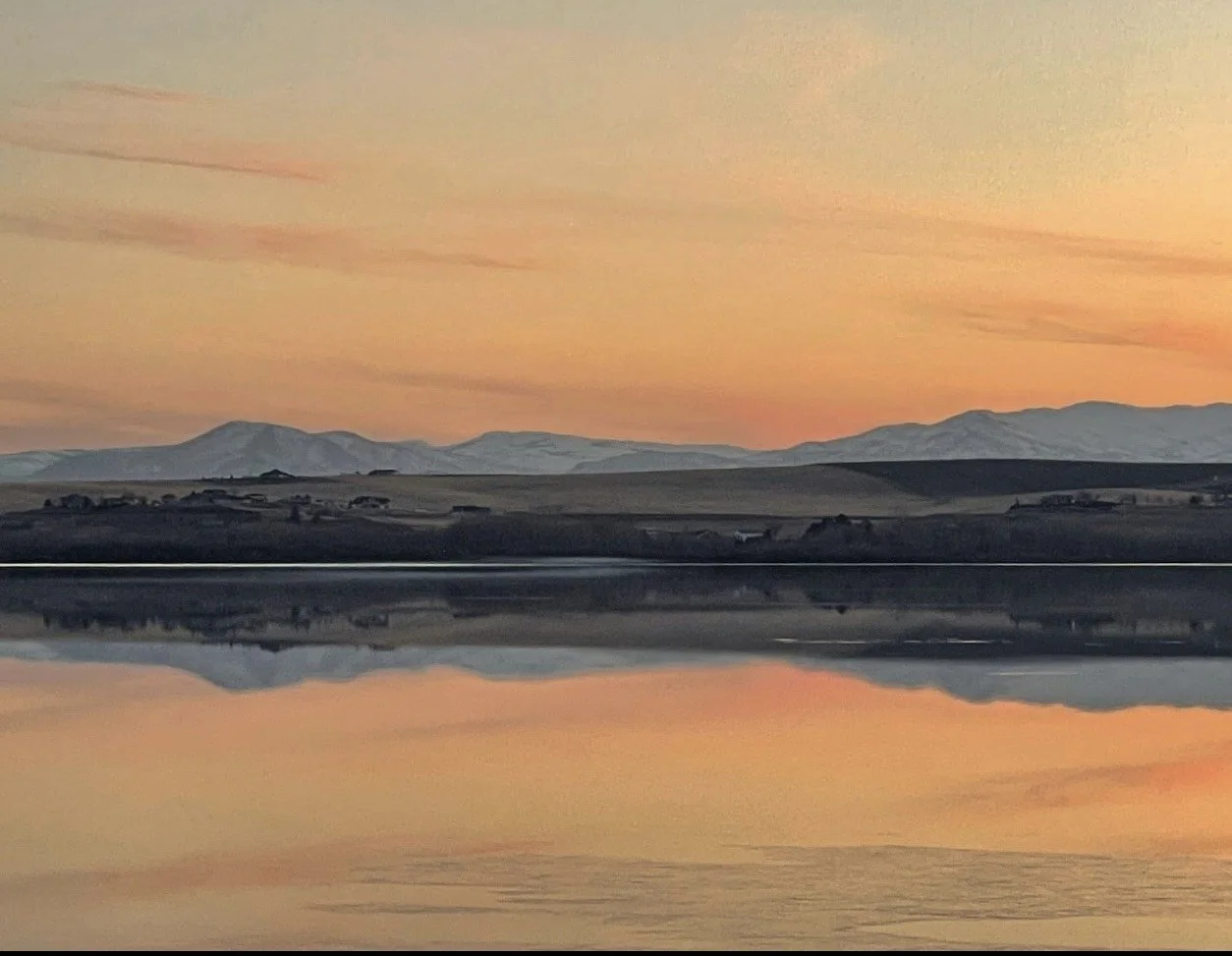A peaceful landscape of rolling hills with snow-capped mountains in the background during sunset, reflected on a calm body of water in the foreground.