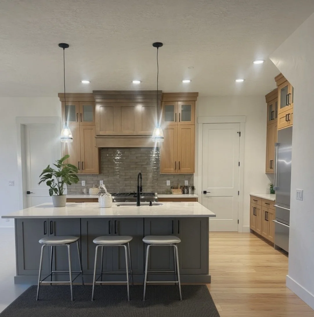 Modern kitchen with oversized island, wooden cabinetry, brick backsplash, black faucet, pendant lights, houseplant, and stainless steel appliances.