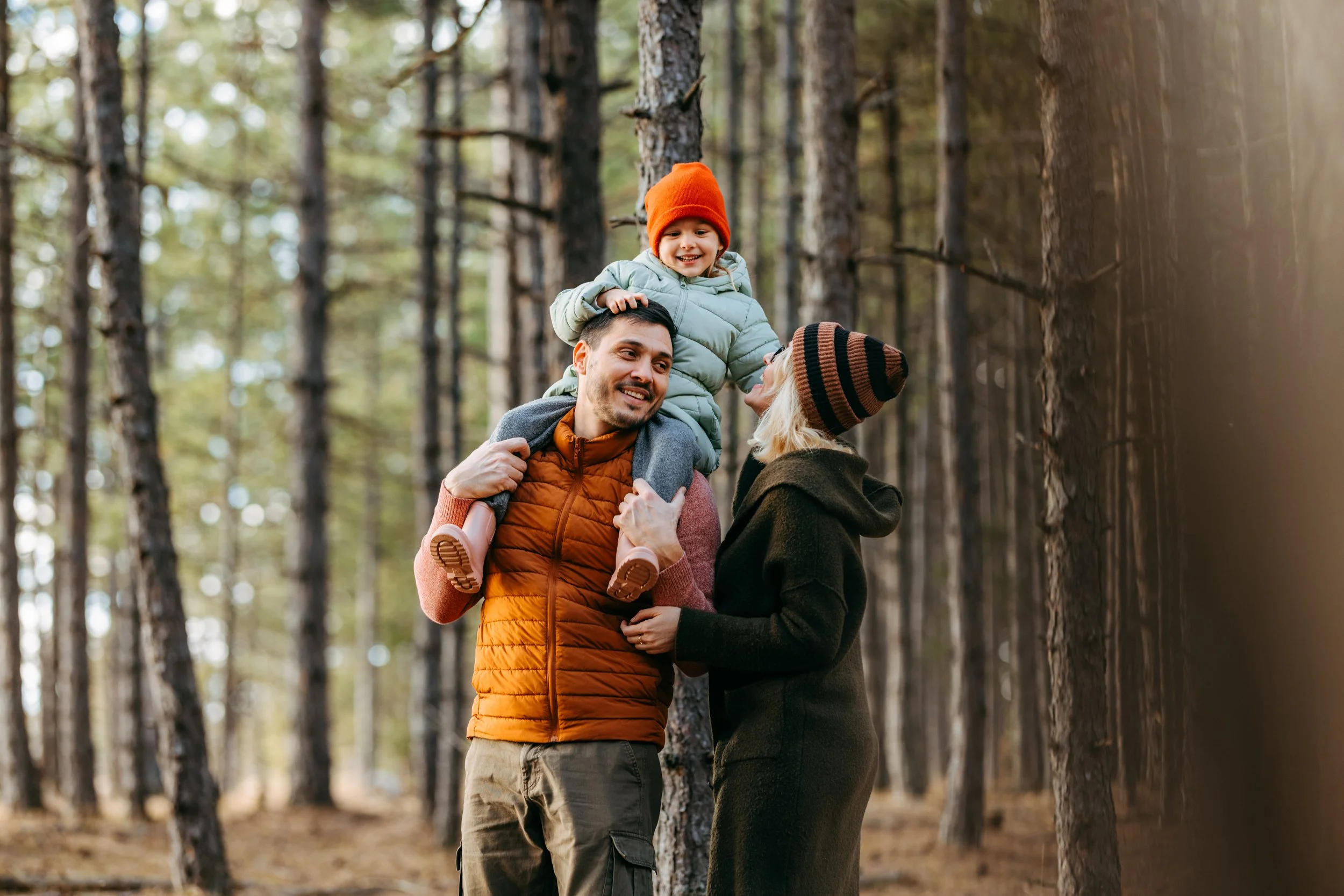 Family in a forest, with a man carrying a smiling young girl on his shoulders, while a woman looks at them affectionately.