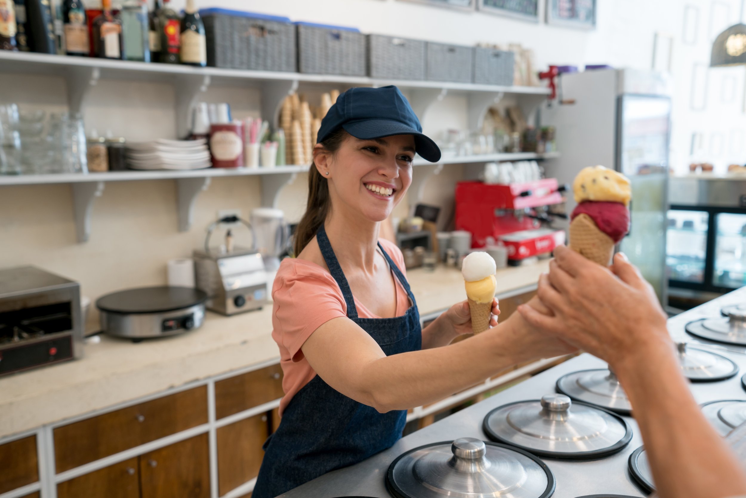 A smiling woman working at an ice cream shop, handing a cone to a customer. The woman is wearing a navy apron and cap, surrounded by kitchen equipment and shelves with supplies.