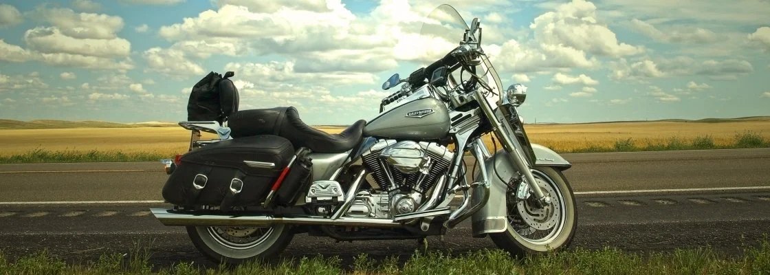 A silver and black touring motorcycle parked on the side of a rural road with open fields and a partly cloudy sky in the background.