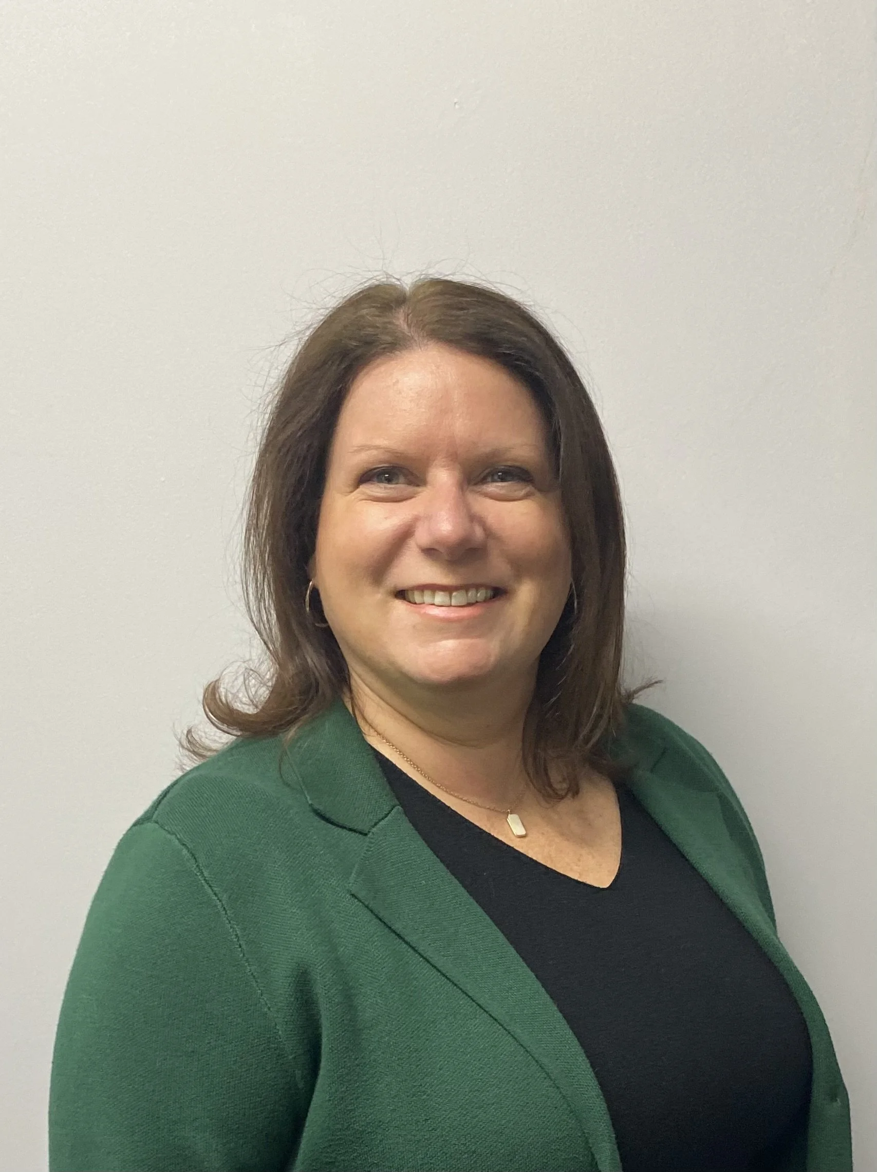 A woman with shoulder-length brown hair, wearing a green blazer over a black top, smiling at the camera against a plain light-colored wall.