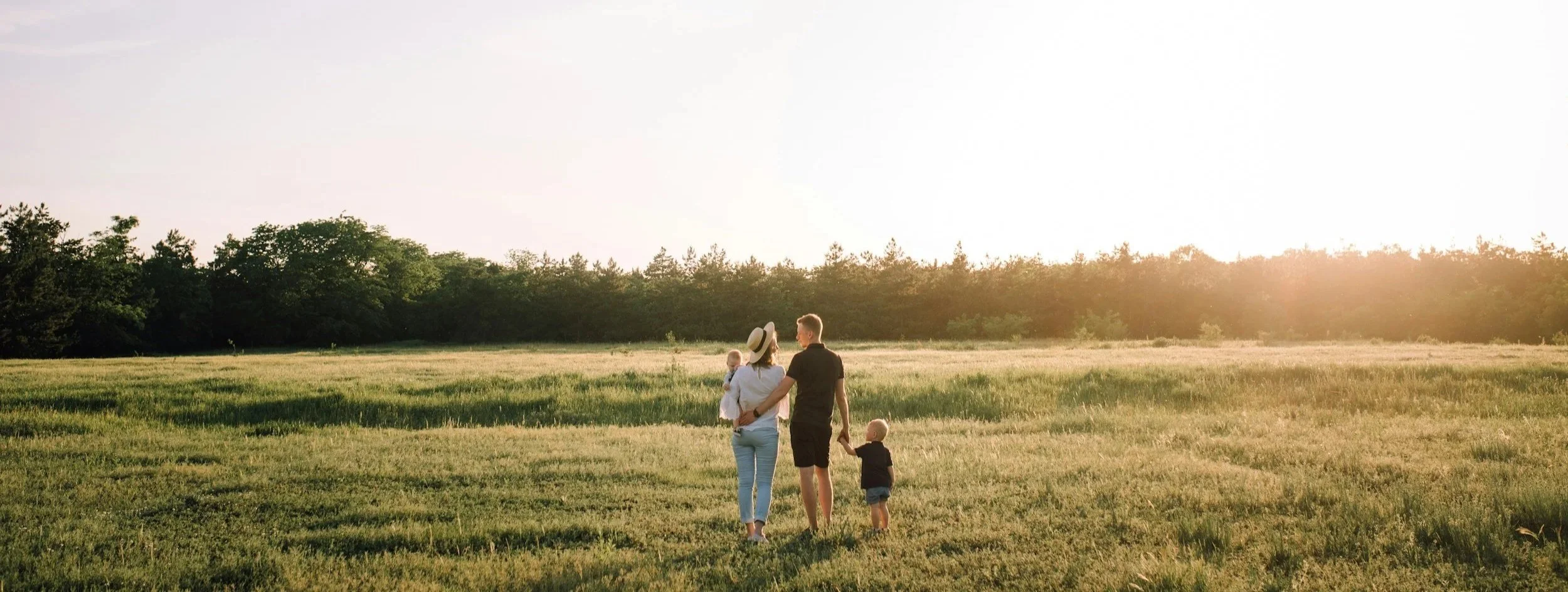 A family of four walking in a grassy field during sunset, with trees in the background.