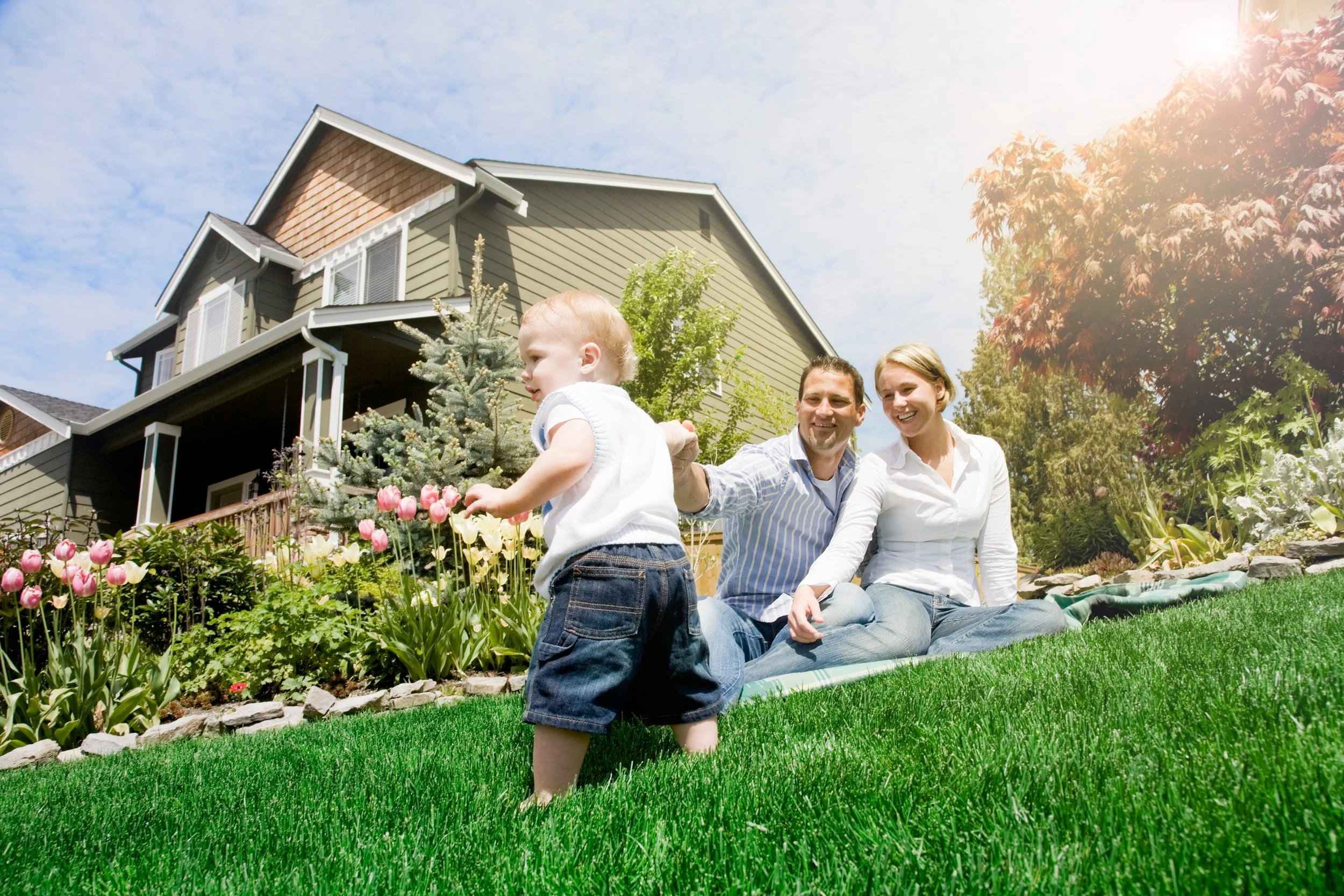A family with a toddler sitting on a blanket on a lawn in front of a house with a garden and trees, under a bright sky with the sun shining.