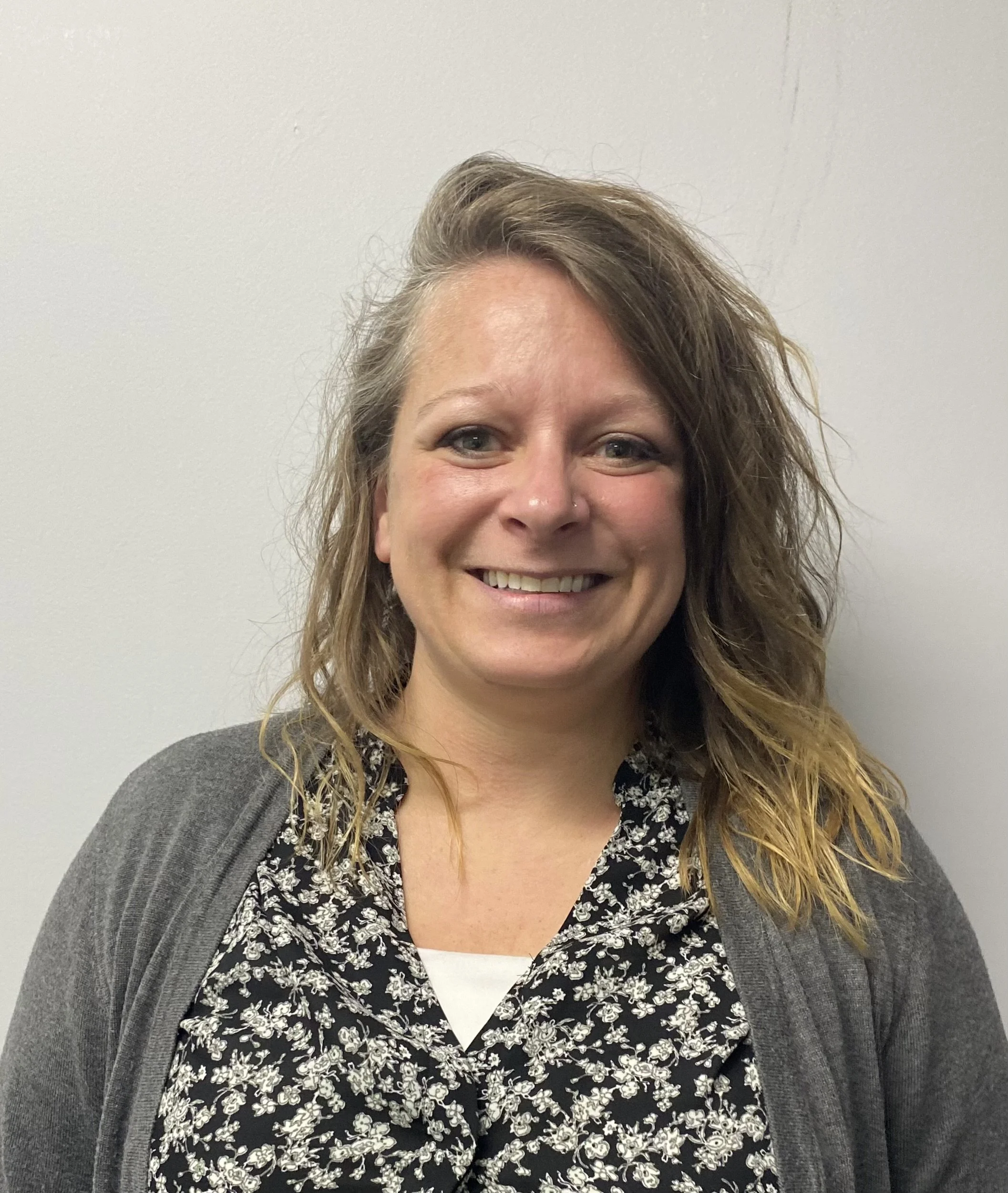 A woman smiling, with wavy brown hair, wearing a black and white floral top and a gray cardigan, standing against a plain white wall.