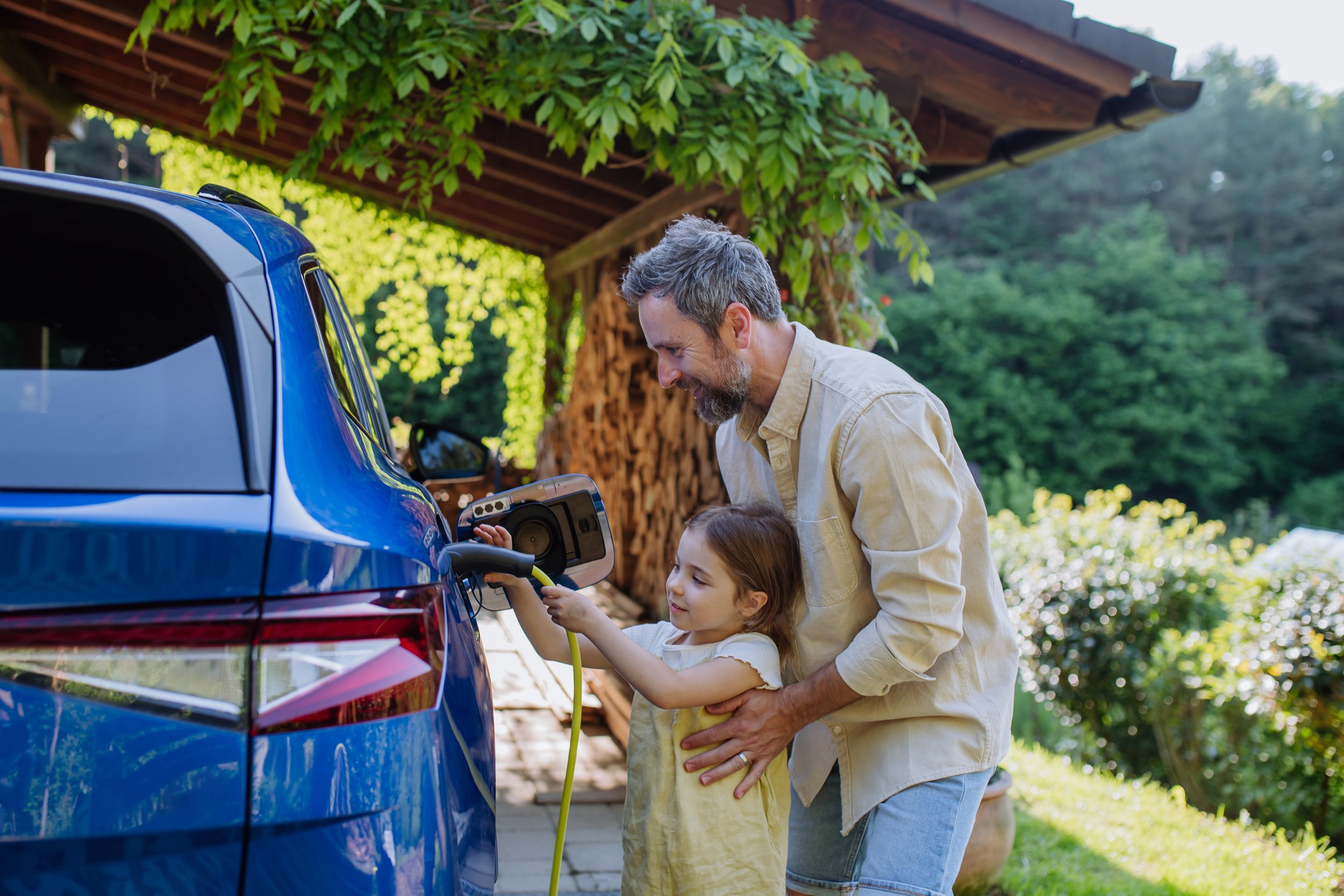 A man and a young girl are charging a blue electric car outdoors in a green, wooded area.